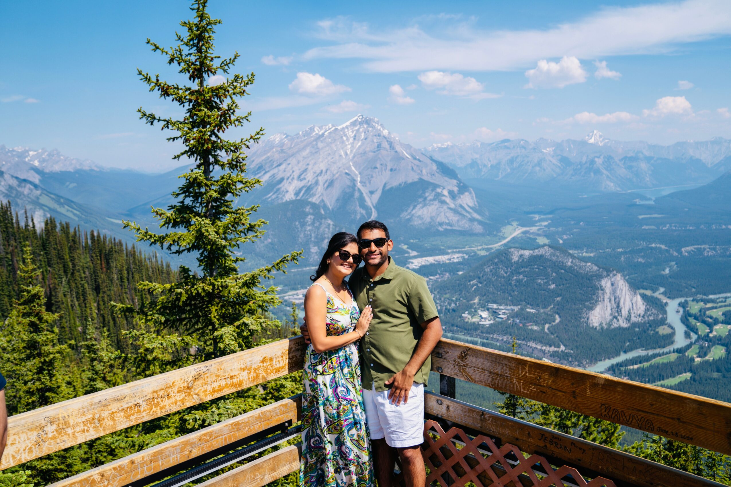 A couple stands on a wooden balcony overlooking mountains and a valley with a river, smiling at the camera.
