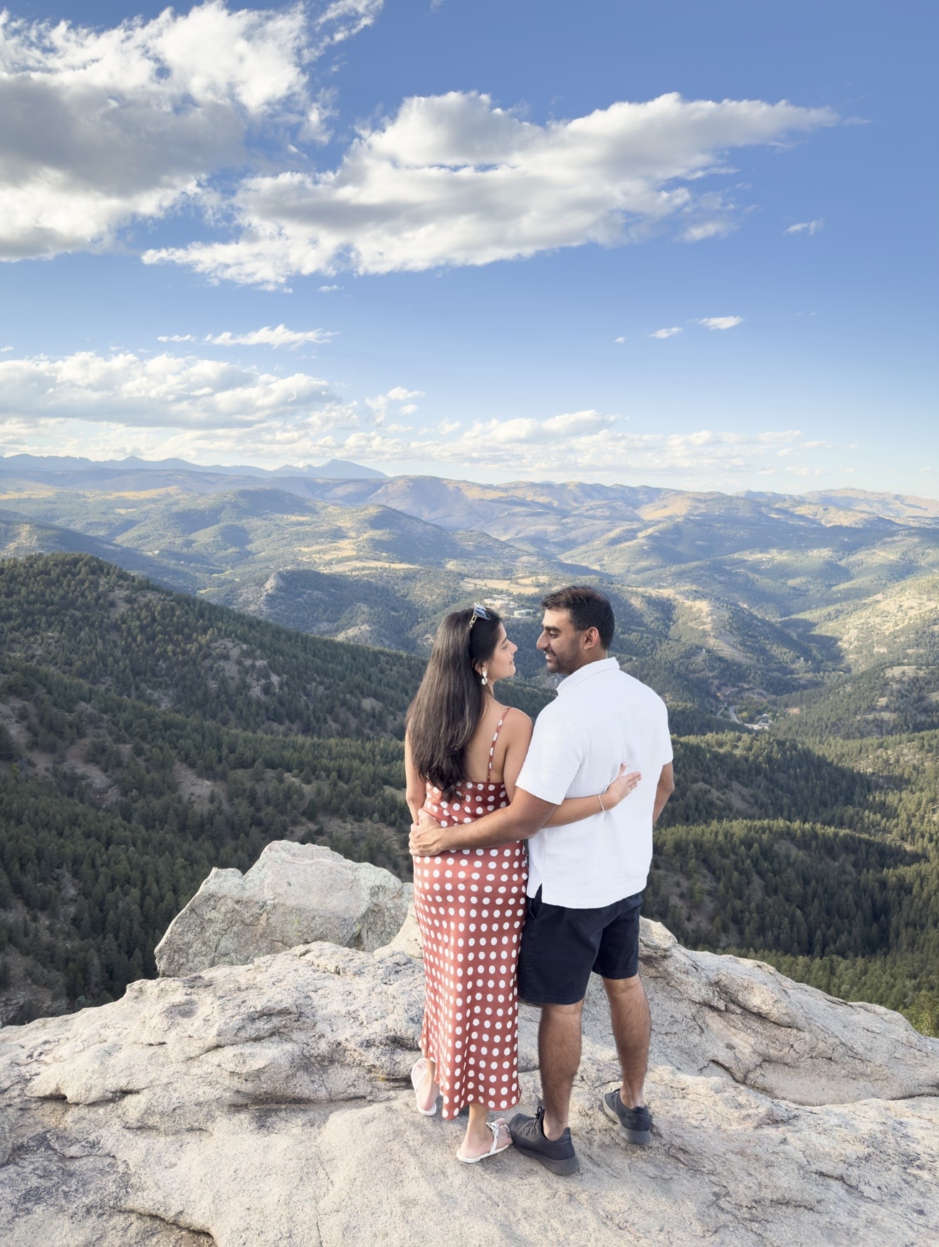 A couple stands on a rocky ledge overlooking mountains and a valley under a partly cloudy sky.