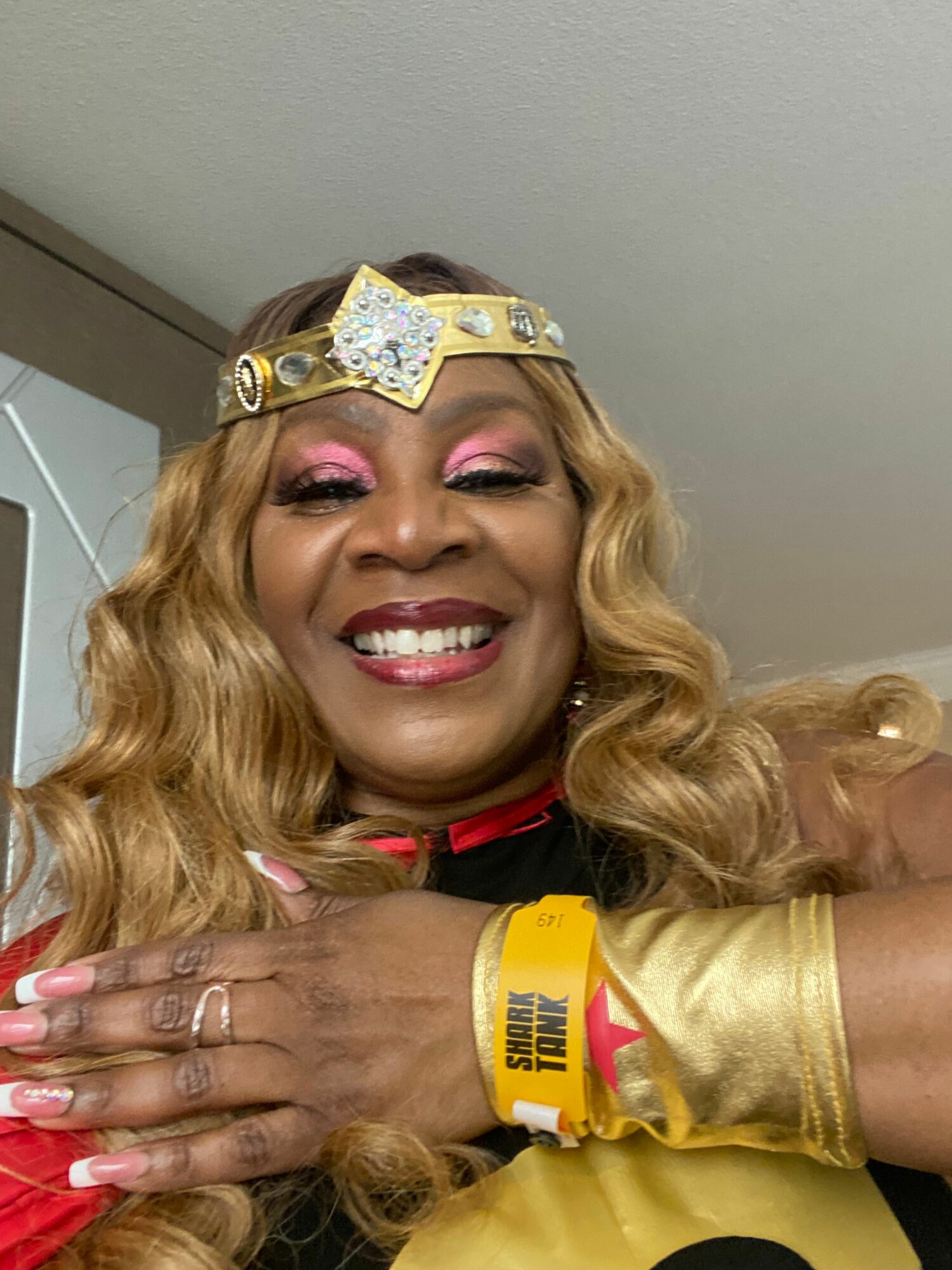 Person with long blonde hair, wearing a crown, smiling, with pink makeup, and a yellow wristband, lying down.