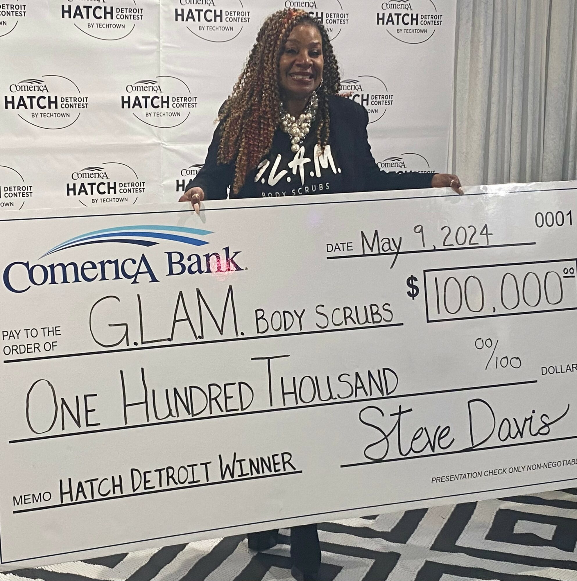 Woman with curly hair and a pearl necklace holding a large check for $100,000 at an event, smiling.