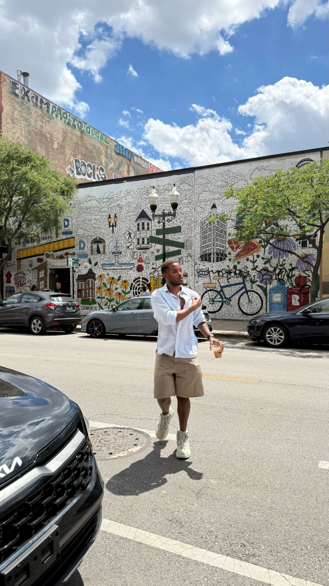 Man standing on street with colorful mural wall, cars parked, trees, and blue sky with clouds in background.