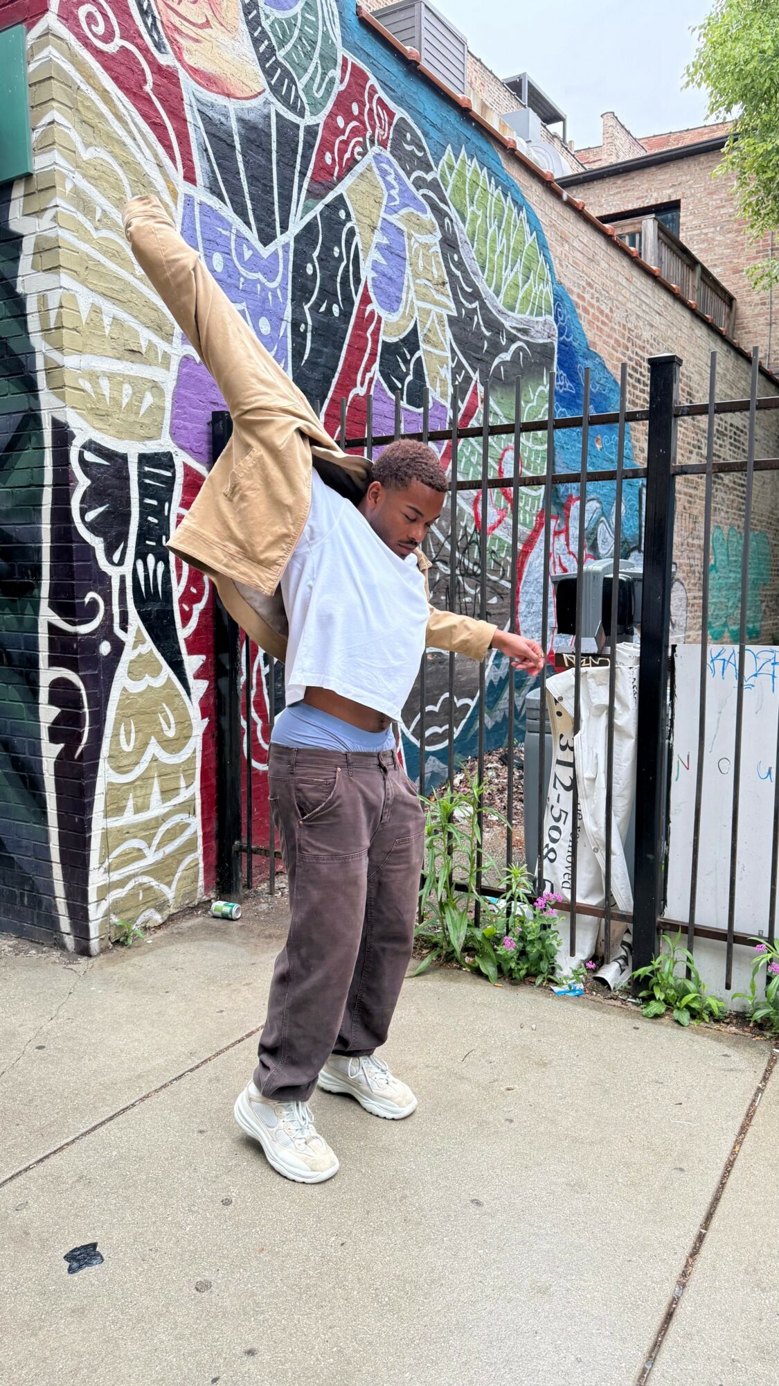 Person dancing outdoors in front of a colorful mural, arms raised, wearing a white shirt, brown pants, and white shoes.