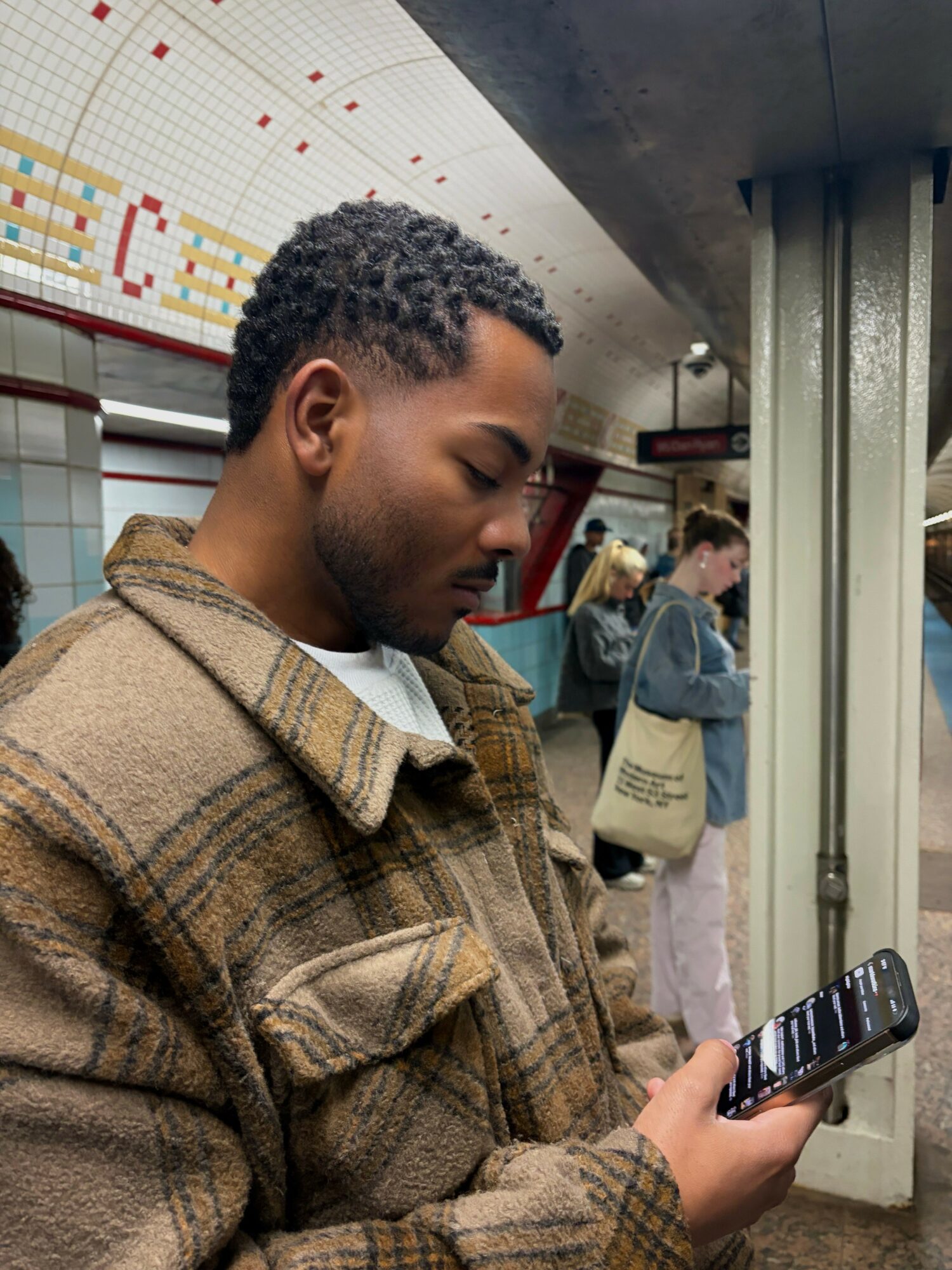 Man with short curly hair looking at a smartphone in a subway station, with other people in the background.