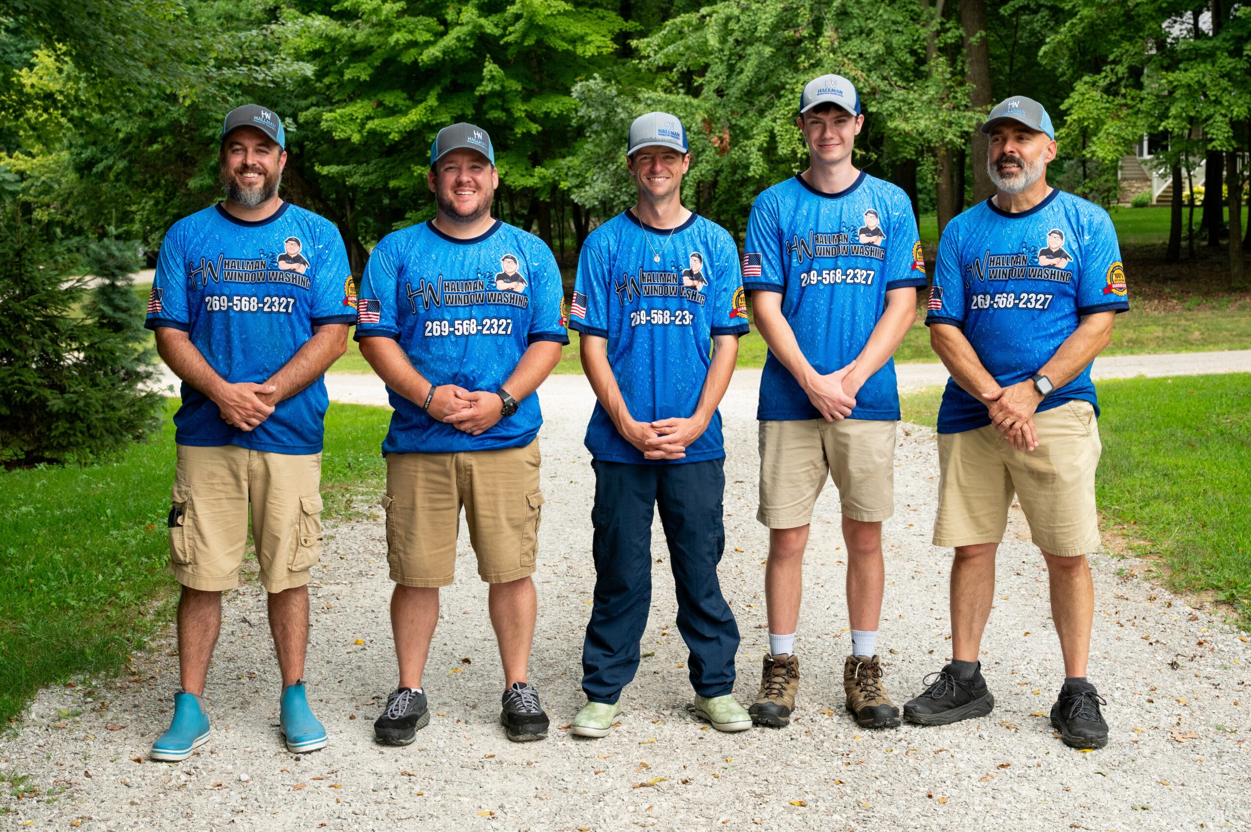 Five people stand outdoors in a line, wearing matching blue shirts and khaki shorts, with trees in the background.