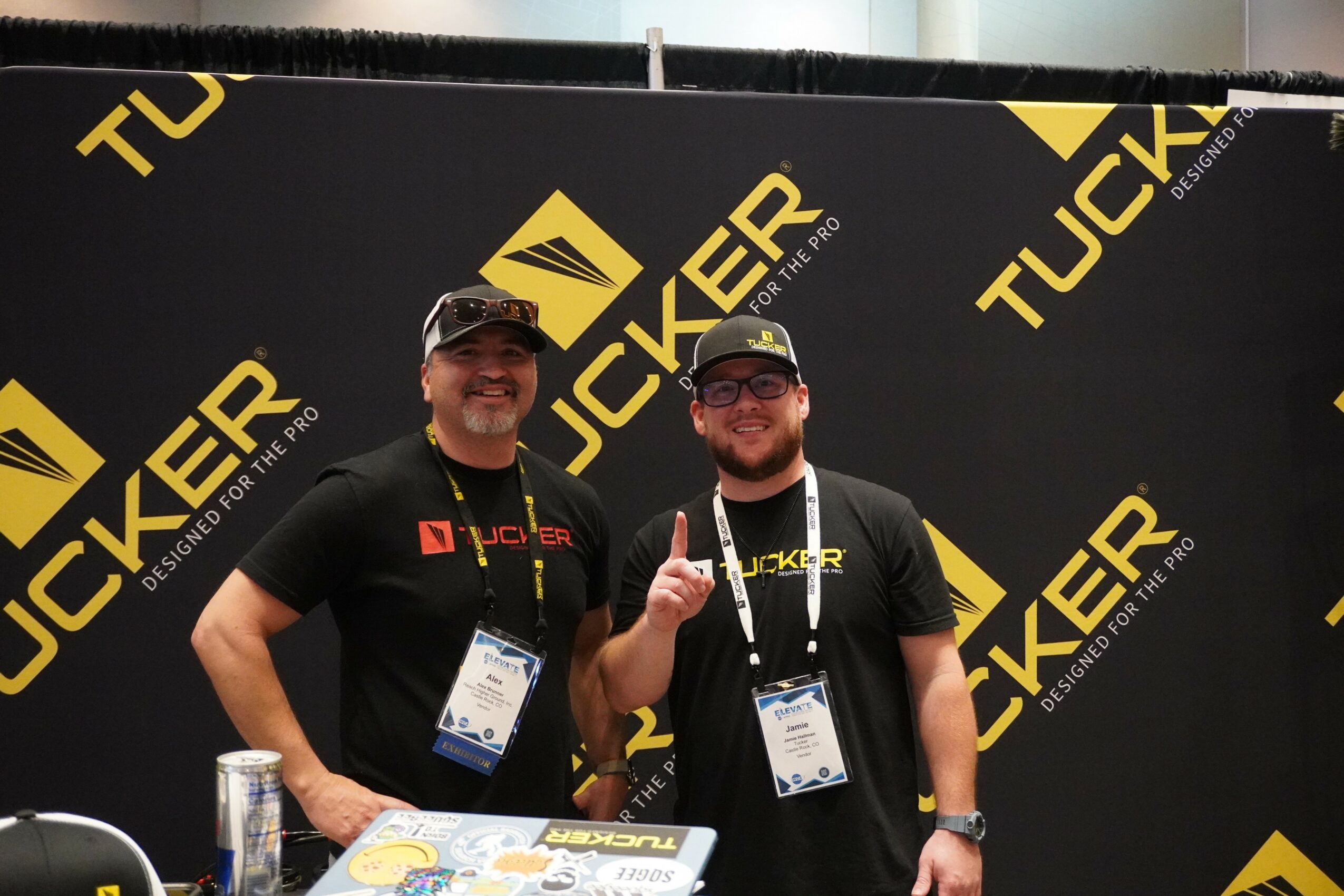 Two men wearing black TuckER shirts and caps, standing in front of a black and yellow TuckER backdrop, smiling and giving thumbs up.
