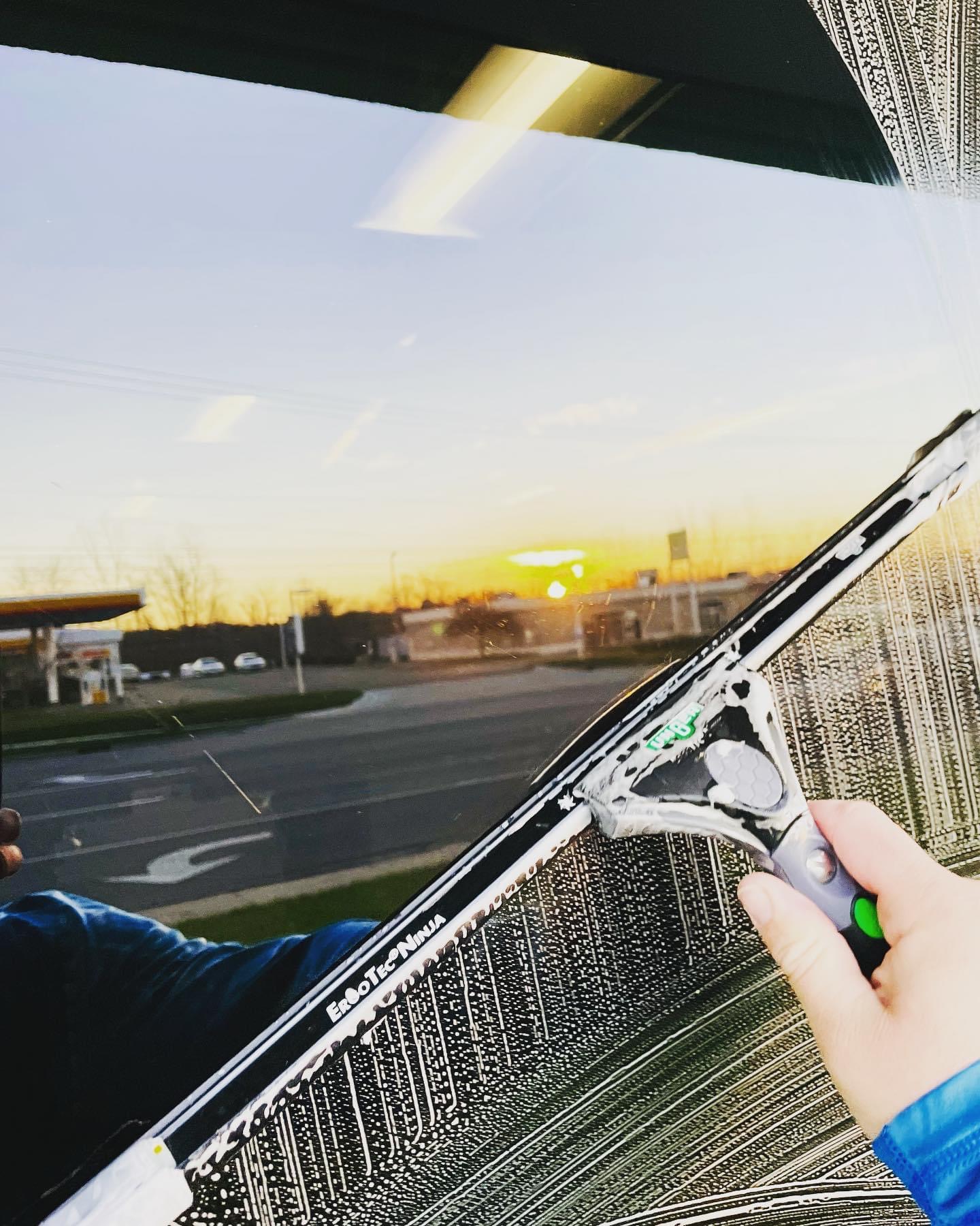 Person cleaning a window with a squeegee, view of a parking lot and sunset outside.