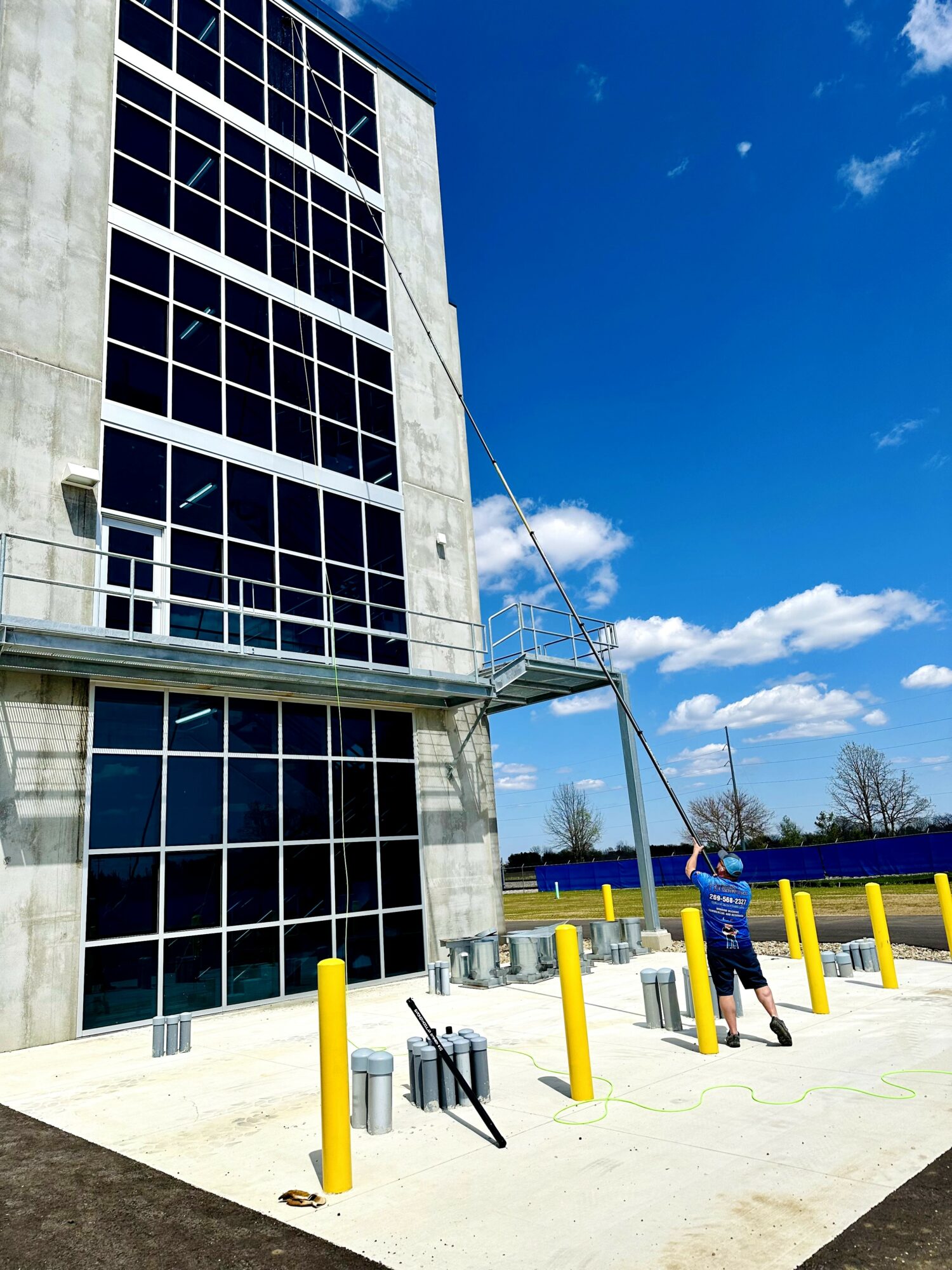 Person cleaning windows on a tall building with a blue sky background, yellow safety posts nearby.