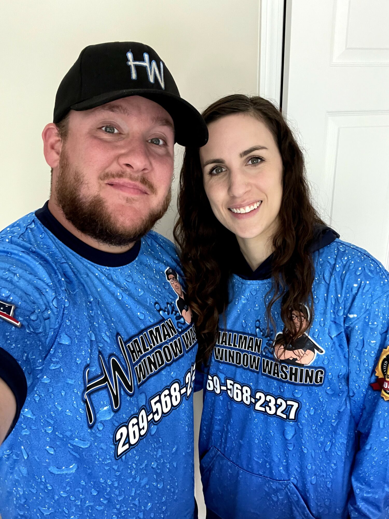 Two people wearing blue sports jerseys with text and phone number, smiling indoors against a white wall.