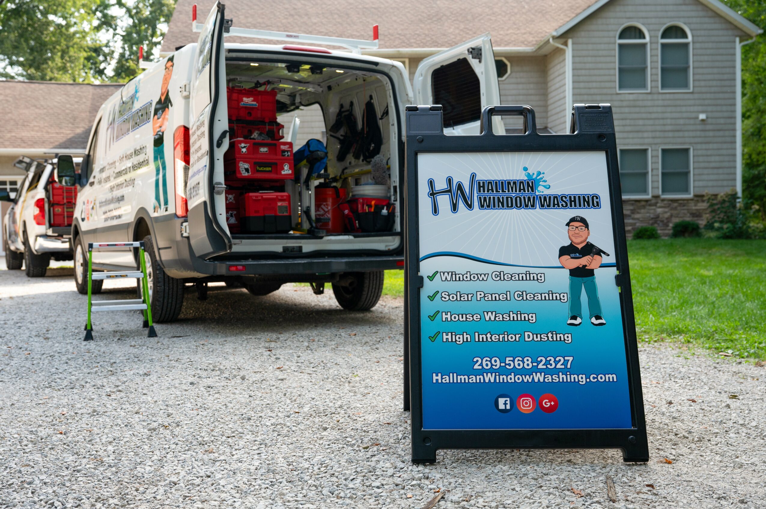 Van with open rear doors parked on driveway, advertising window washing services on a signboard, house in background.