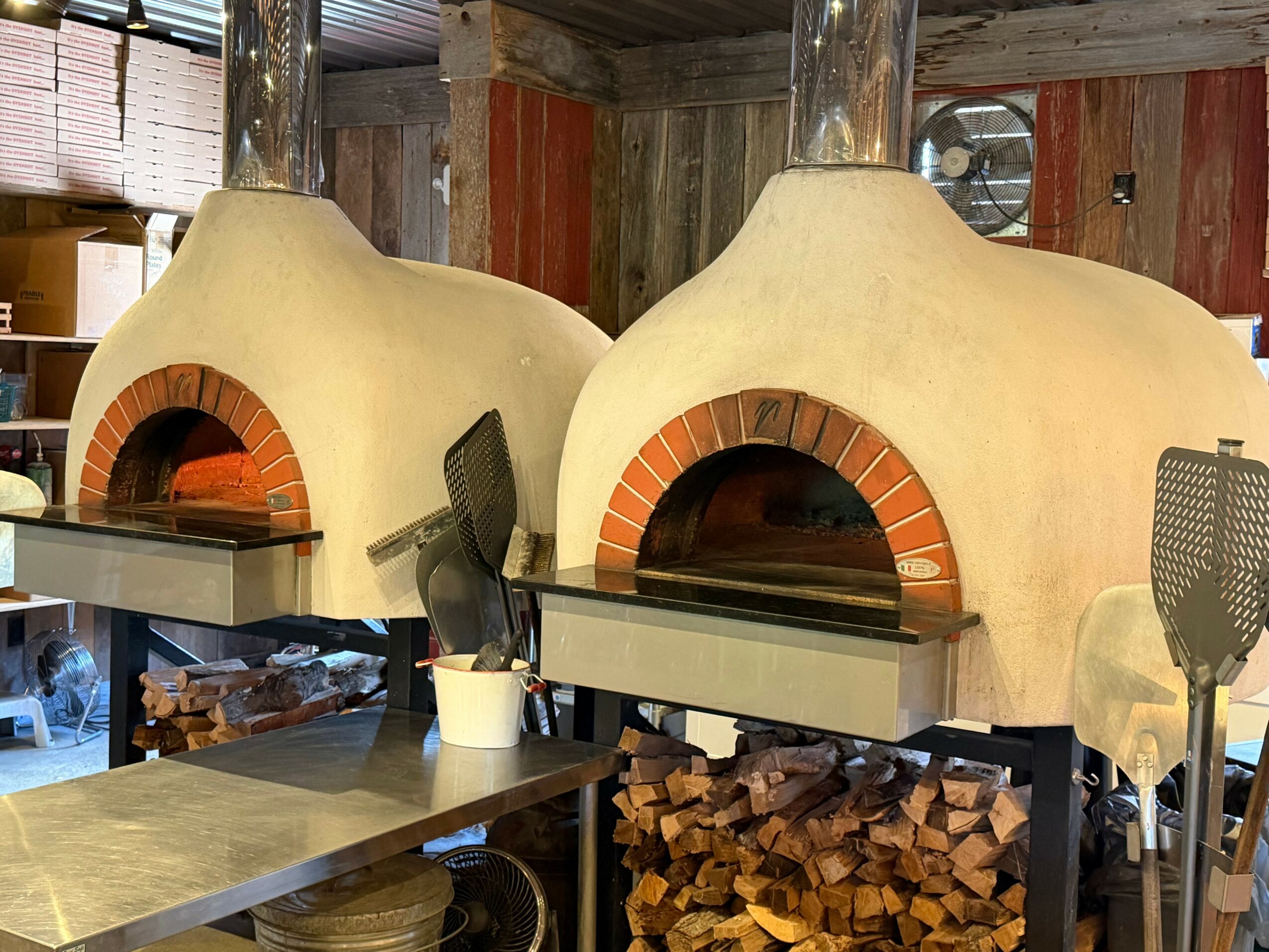 Two wood-fired pizza ovens with brick arches, surrounded by kitchen tools and firewood, in a rustic setting.