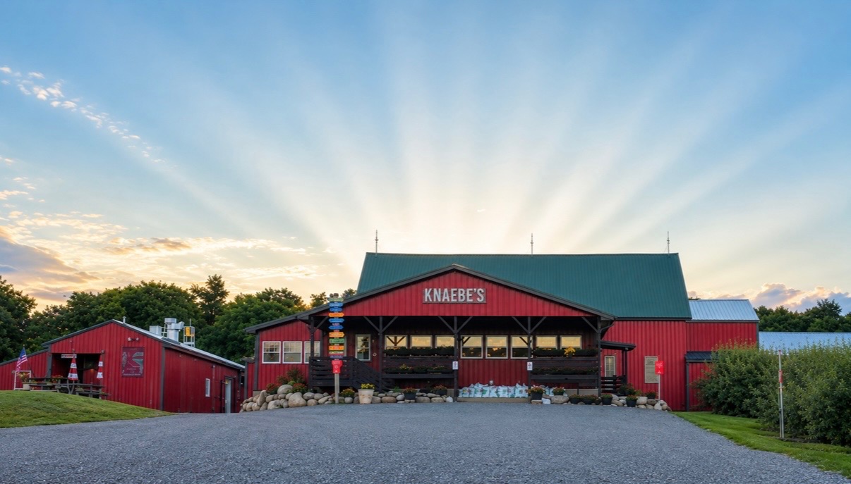 Red barn-style building with a green roof and a sign reading 'KNAEBES' under a bright sky with sun rays.