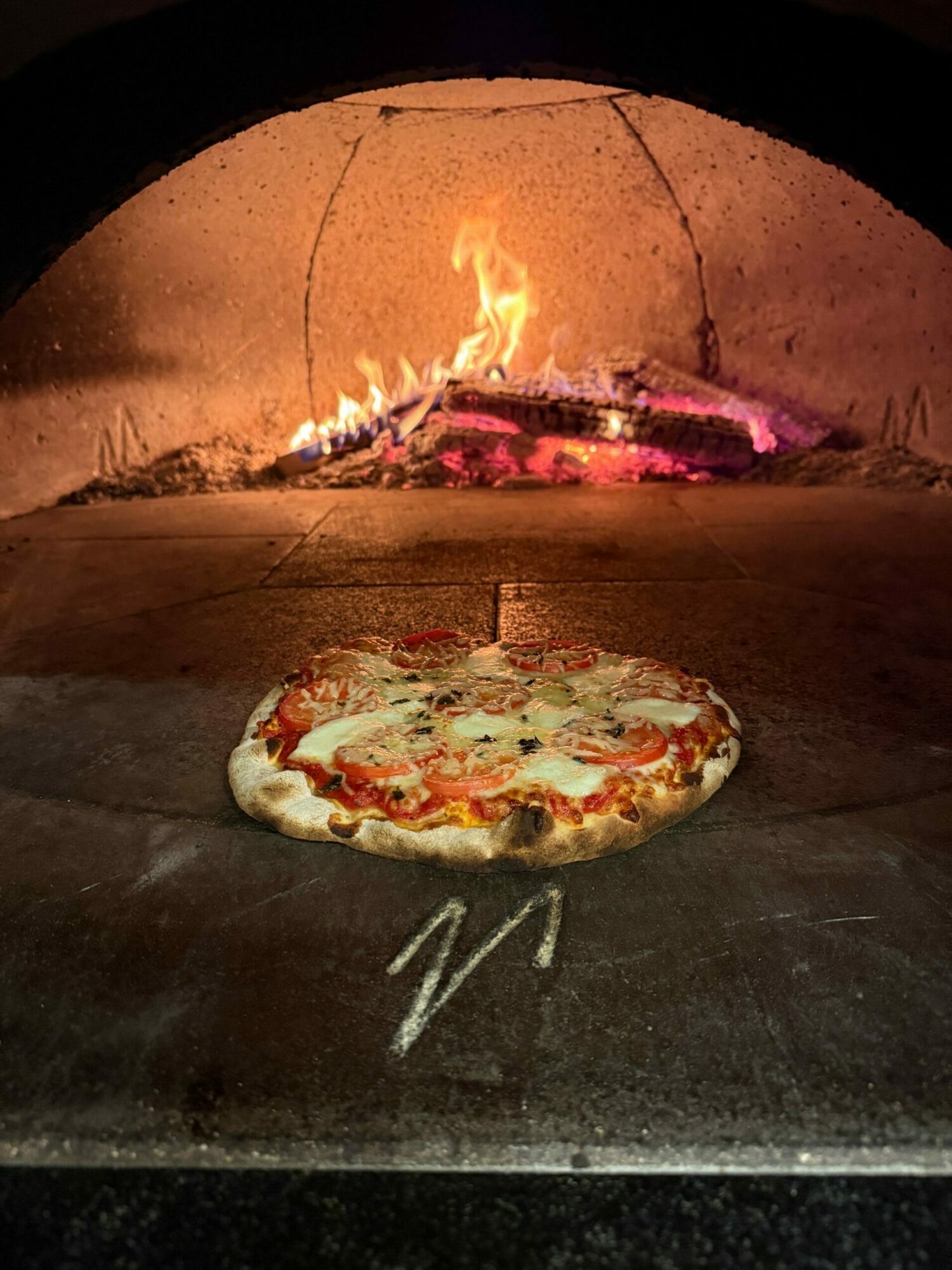 Pizza on a stone surface with a fire in the background, glowing embers, and a dark setting.