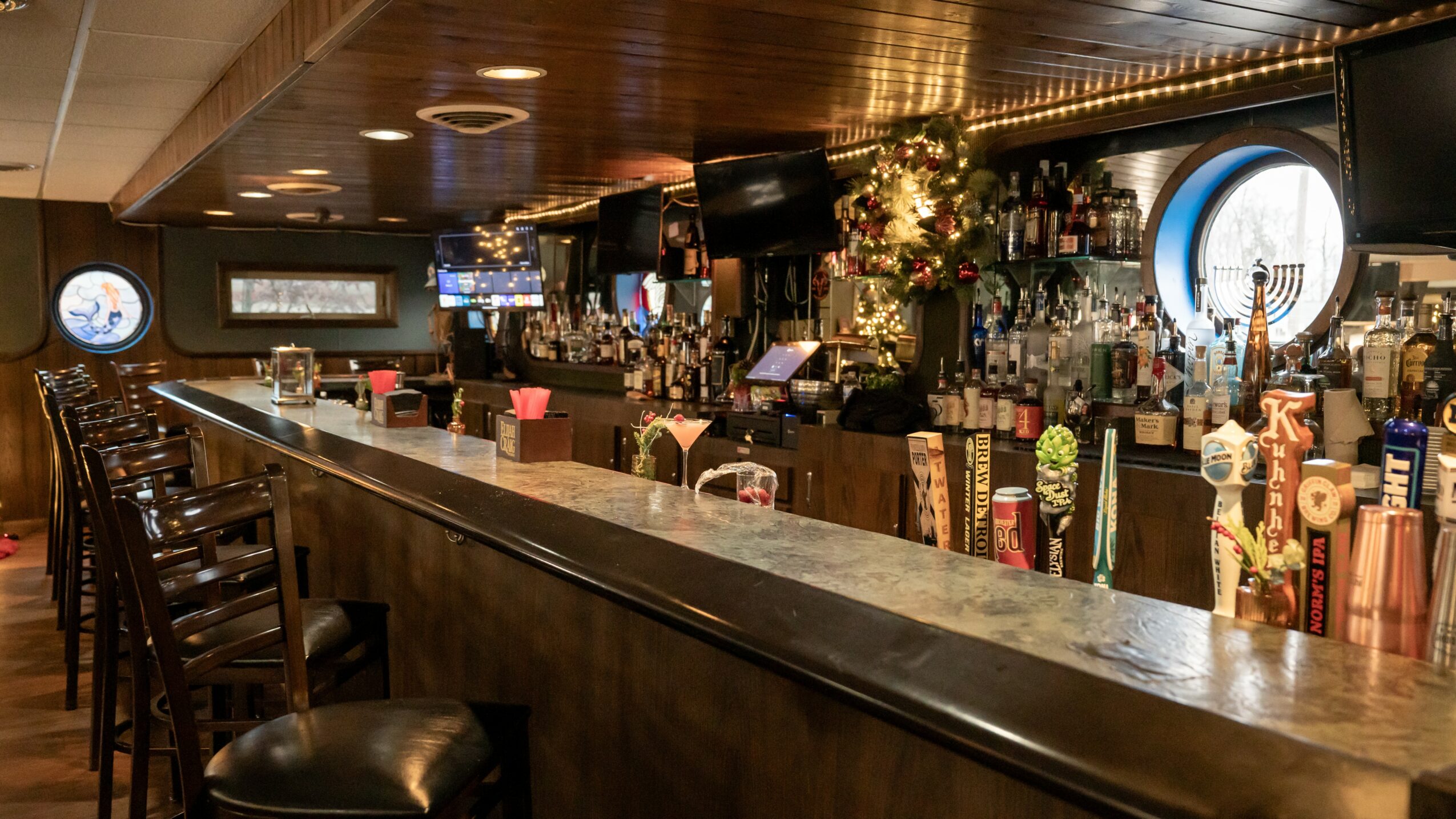 Bar counter with bottles, taps, and seating area, illuminated by warm lighting, with a circular window in the background.
