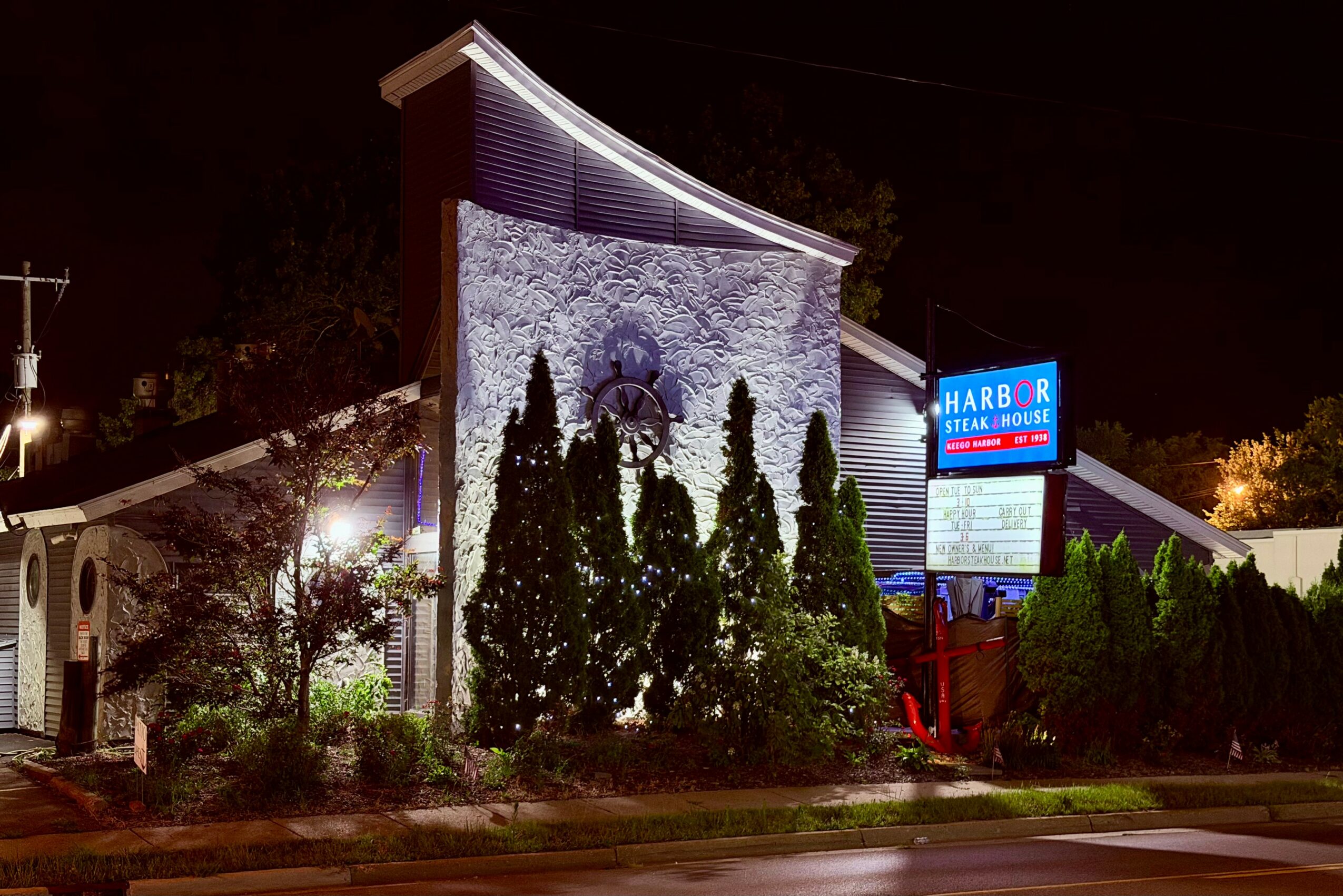 Night view of a building with a curved roof, stone facade, surrounded by trees and a lit sign, illuminated against the dark sky.