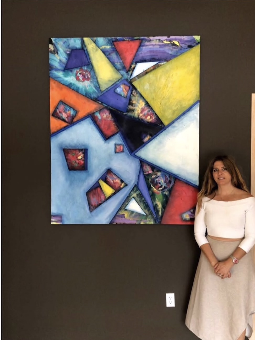 Young woman in white top and beige skirt standing next to colorful abstract painting on dark wall.