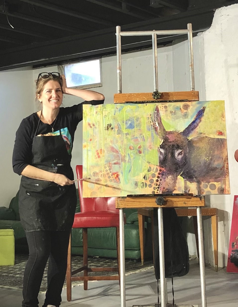 Woman standing next to a colorful painting of a donkey on an easel, smiling and resting her arm on it.