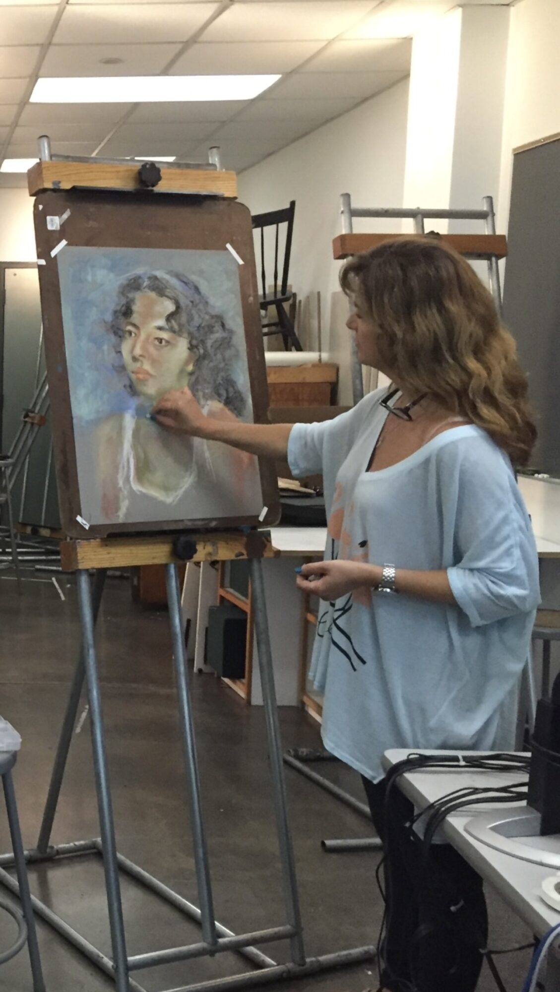 Woman painting a portrait on an easel in an indoor studio, with chairs and equipment around.