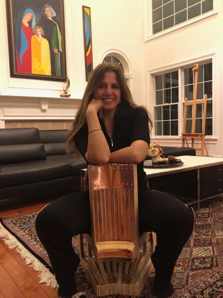 Woman sitting on a chair in a living room, smiling, with artwork and windows in the background.