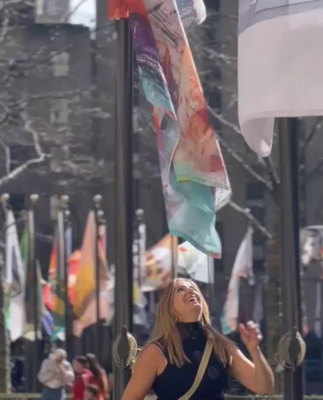 Woman looking up at colorful flags hanging above her outdoors, with trees and buildings in the background.