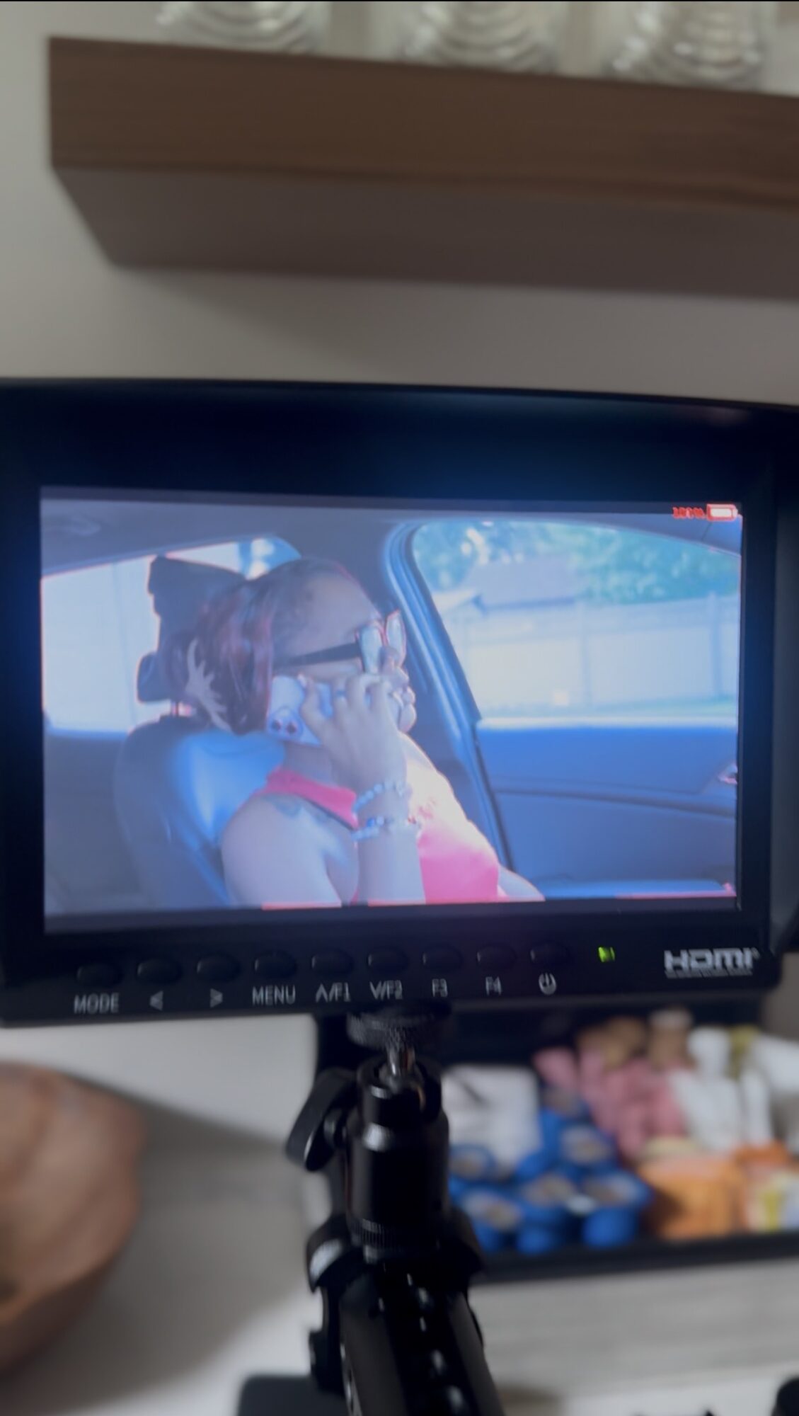 Person with glasses and curly hair sitting in a car, holding a phone to their ear, wearing a red top.