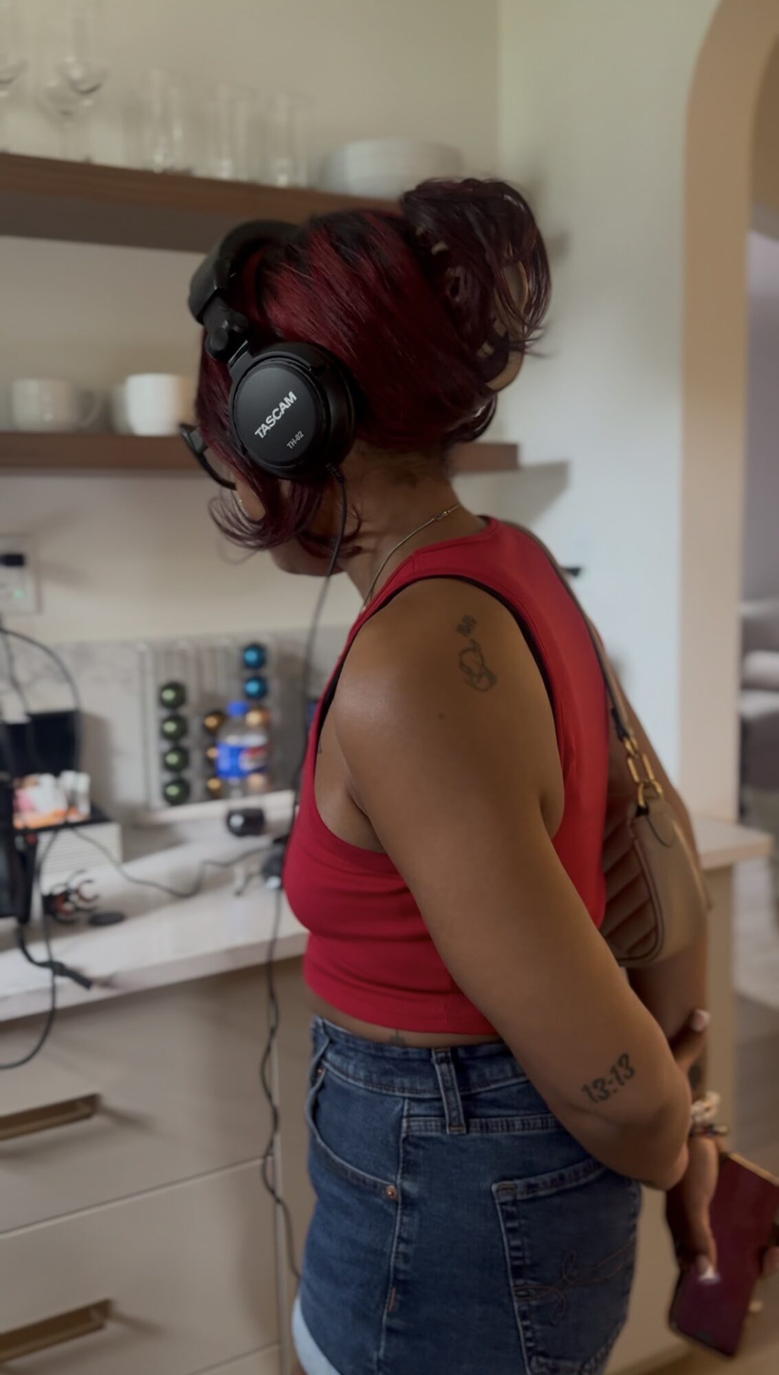 Person with red hair wearing headphones, red sleeveless top, and jeans, standing in a kitchen. Tattoos visible on arm.