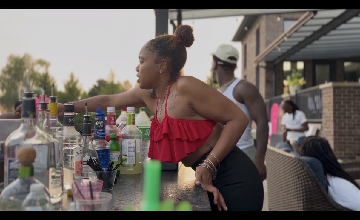Woman reaching for drinks at an outdoor bar with bottles and glasses, people in background, sunny day.