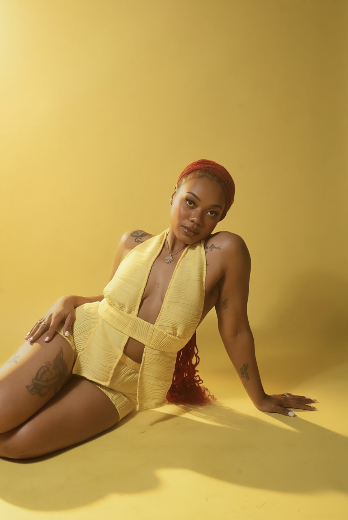 Woman with long red hair in yellow outfit sitting on yellow floor against yellow background, looking at camera.