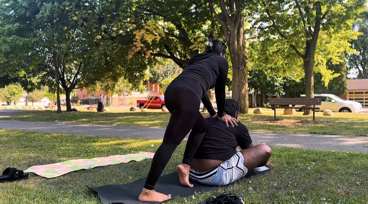 Person receiving a massage outdoors in a park with trees and benches, lying on a mat, another person kneeling beside them.