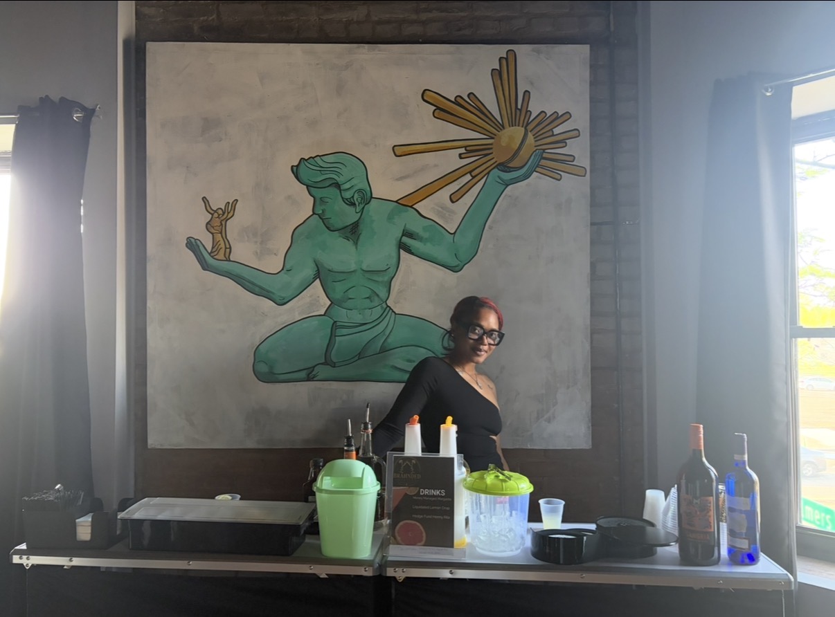 Person with glasses and dark hair standing in front of a large mural of a green figure with multiple arms, holding a small object, inside a room with windows.