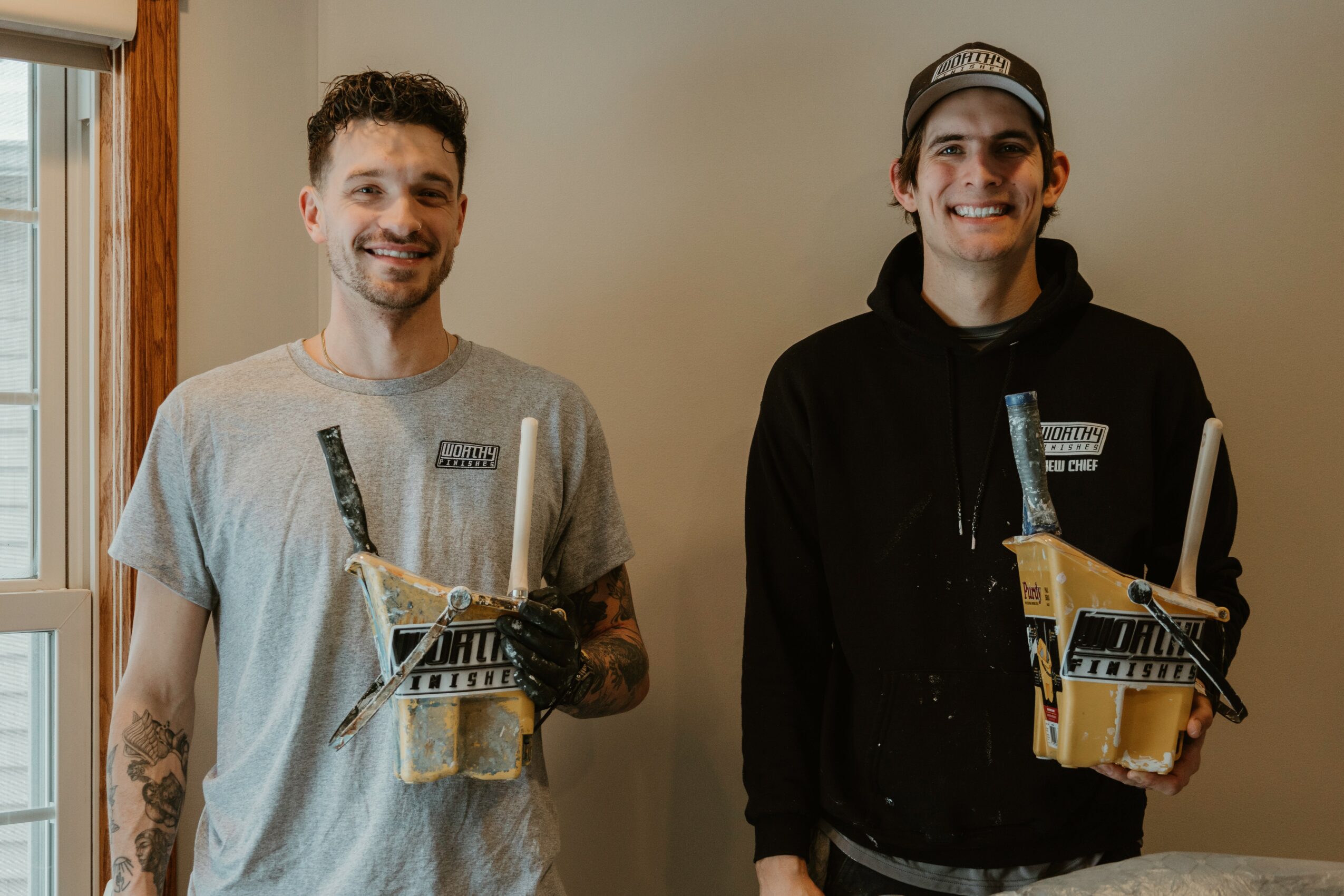 Two smiling young men holding boxes with tools, standing indoors near a window, one wearing a gray t-shirt and the other a black hoodie.
