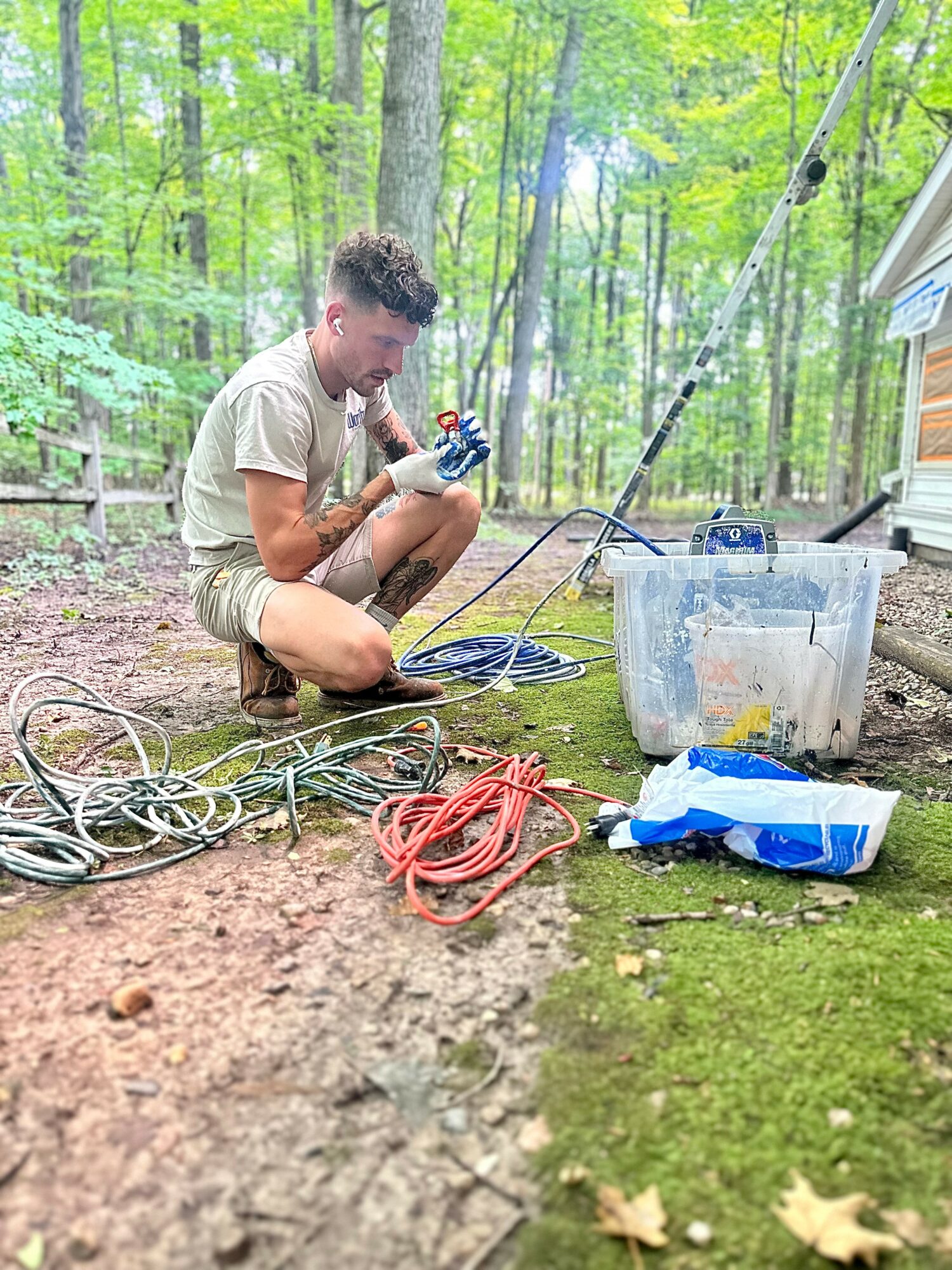 Young man crouching outdoors in a forest, working with electrical cables and tools, with a plastic container and a blue bag nearby.