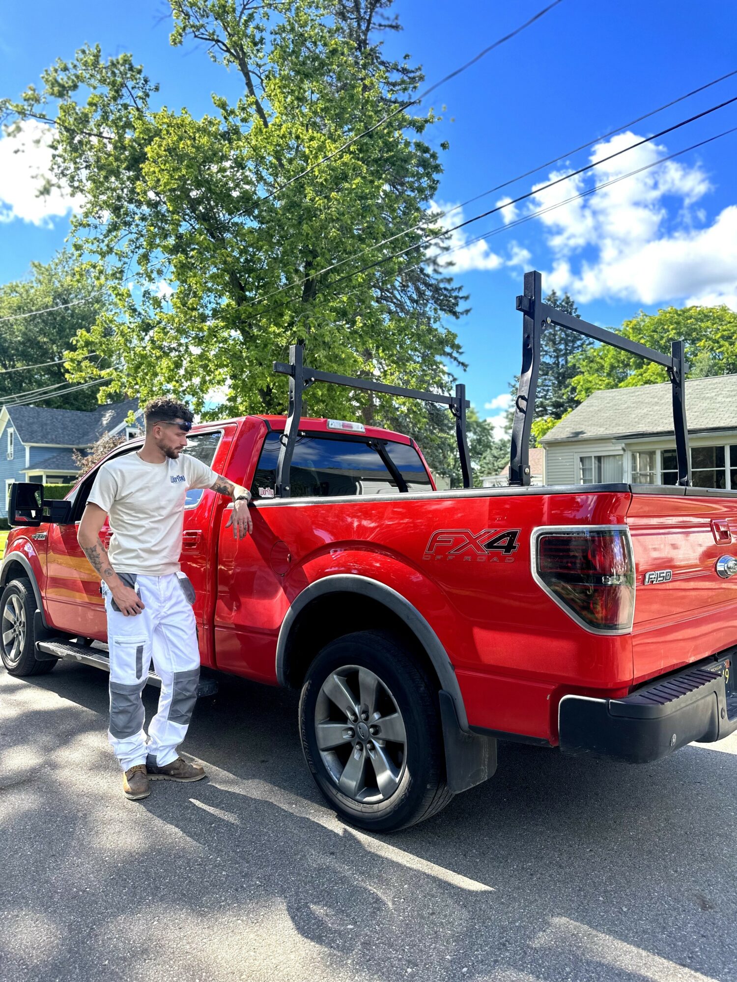 Man in white shirt and jeans standing next to a red pickup truck parked on the street, trees and houses in background.