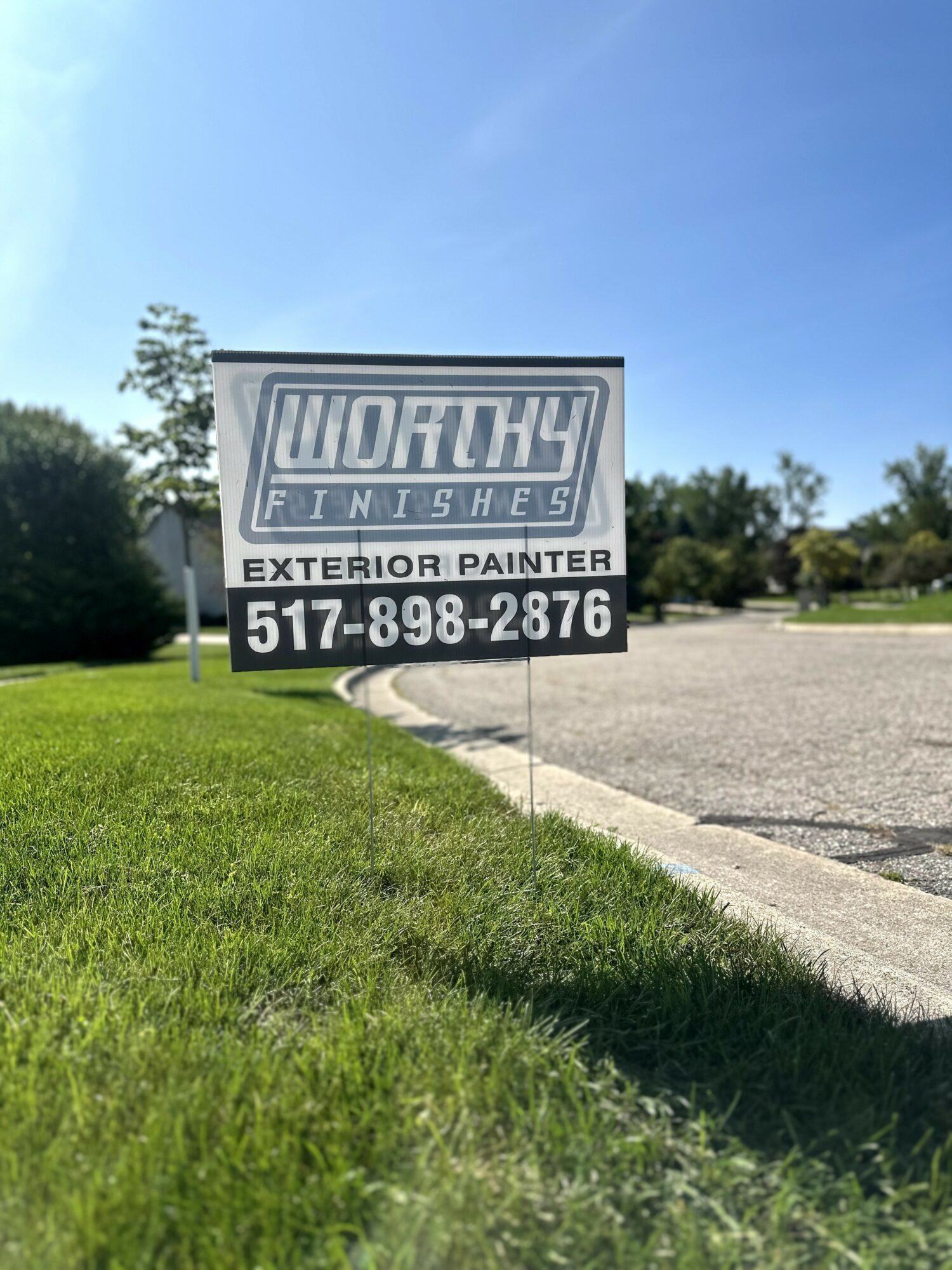 Sign for Works Finishes exterior painter with phone number, on a grassy area near a parking lot, under a clear blue sky.