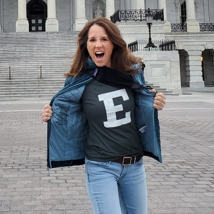 Woman with brown hair, open mouth, wearing a black shirt with a white letter E, blue jeans, and a blue jacket, standing outdoors in front of a building with stairs.