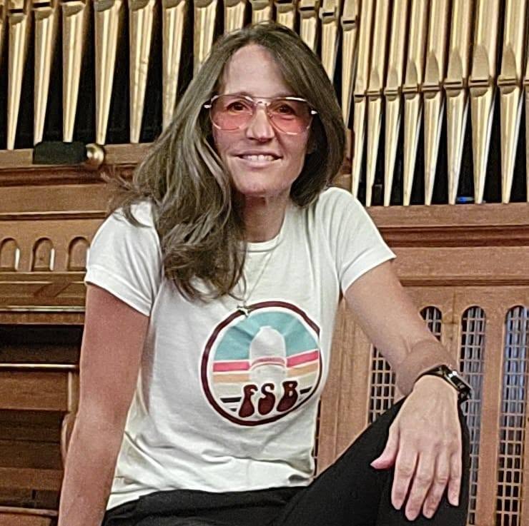 Woman with long hair wearing sunglasses and a white T-shirt, sitting in front of a pipe organ, smiling.