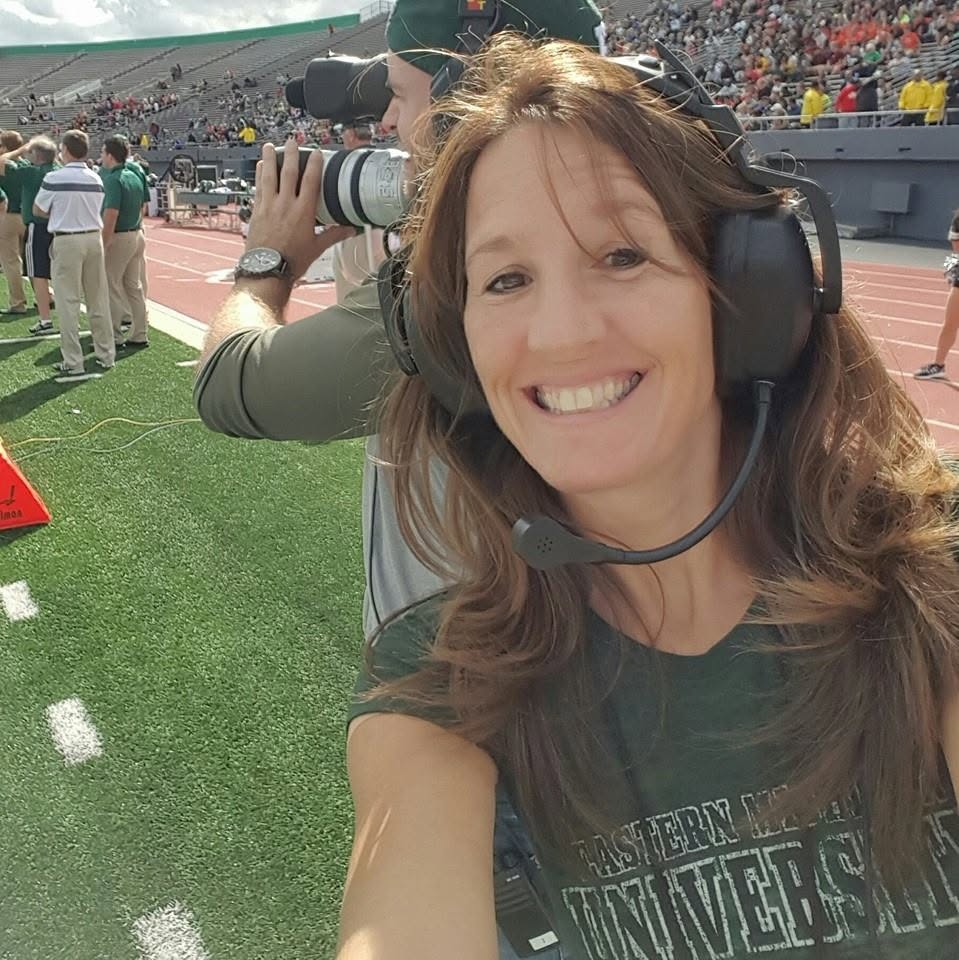 Smiling woman wearing headphones at a sports event, with a camera operator and crowd in background.