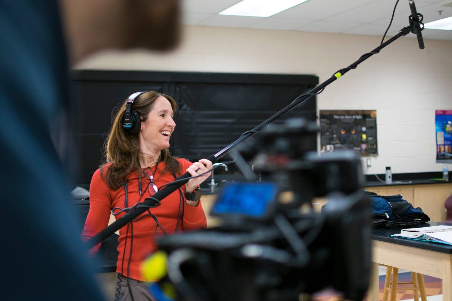 Woman in red shirt wearing headphones speaking into a microphone in a recording studio.