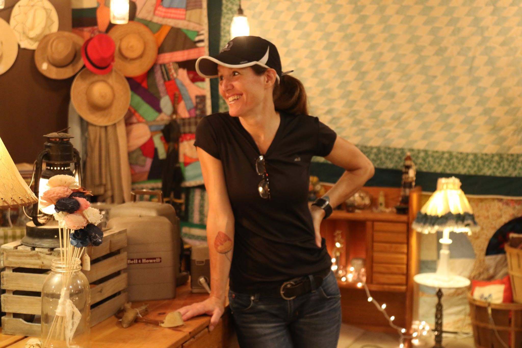 Woman smiling, standing in a decorated room with hats and lamps, wearing a black shirt and cap.