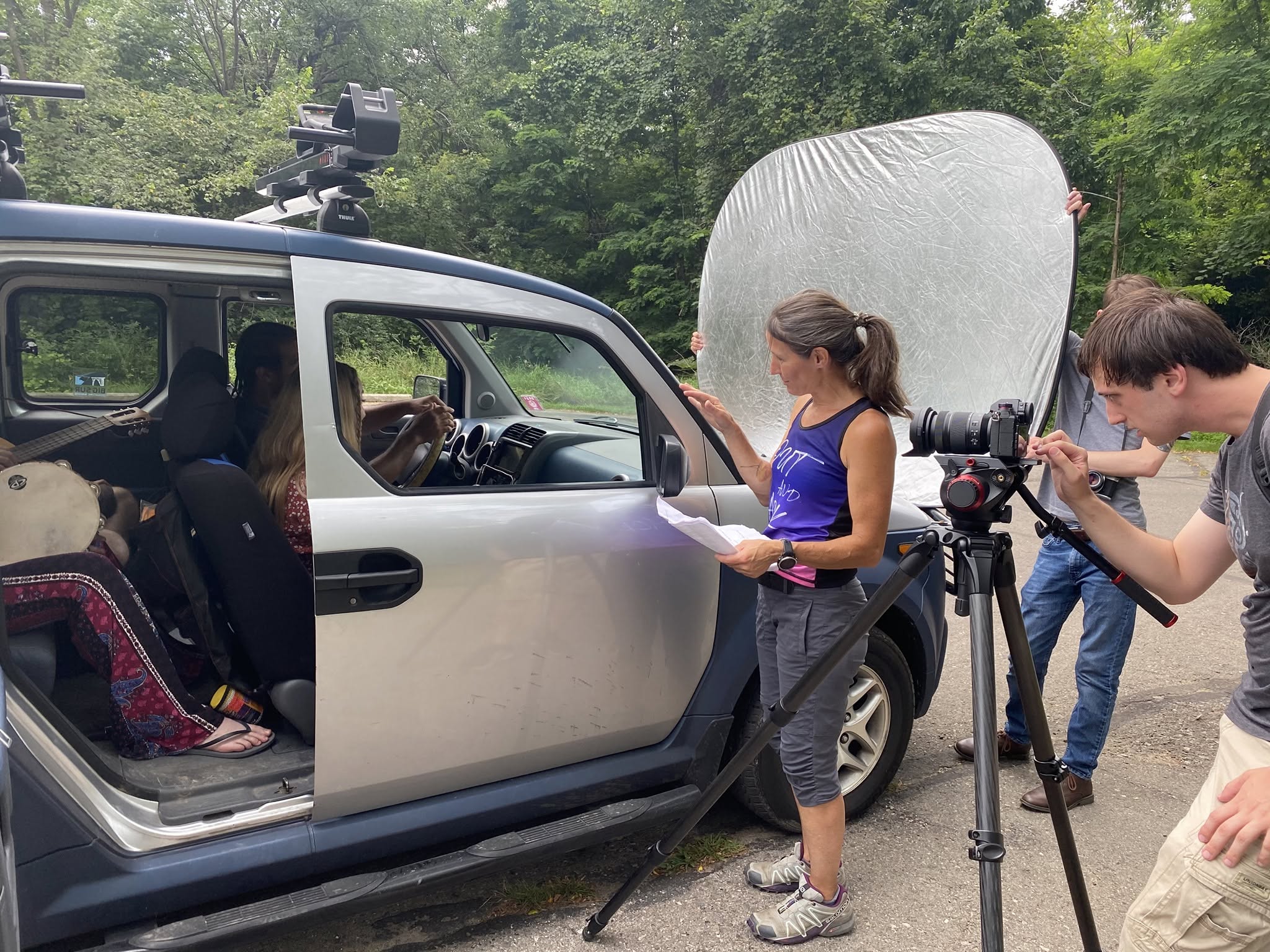 People filming a scene with a camera and reflector outside, with a vehicle and trees in the background.