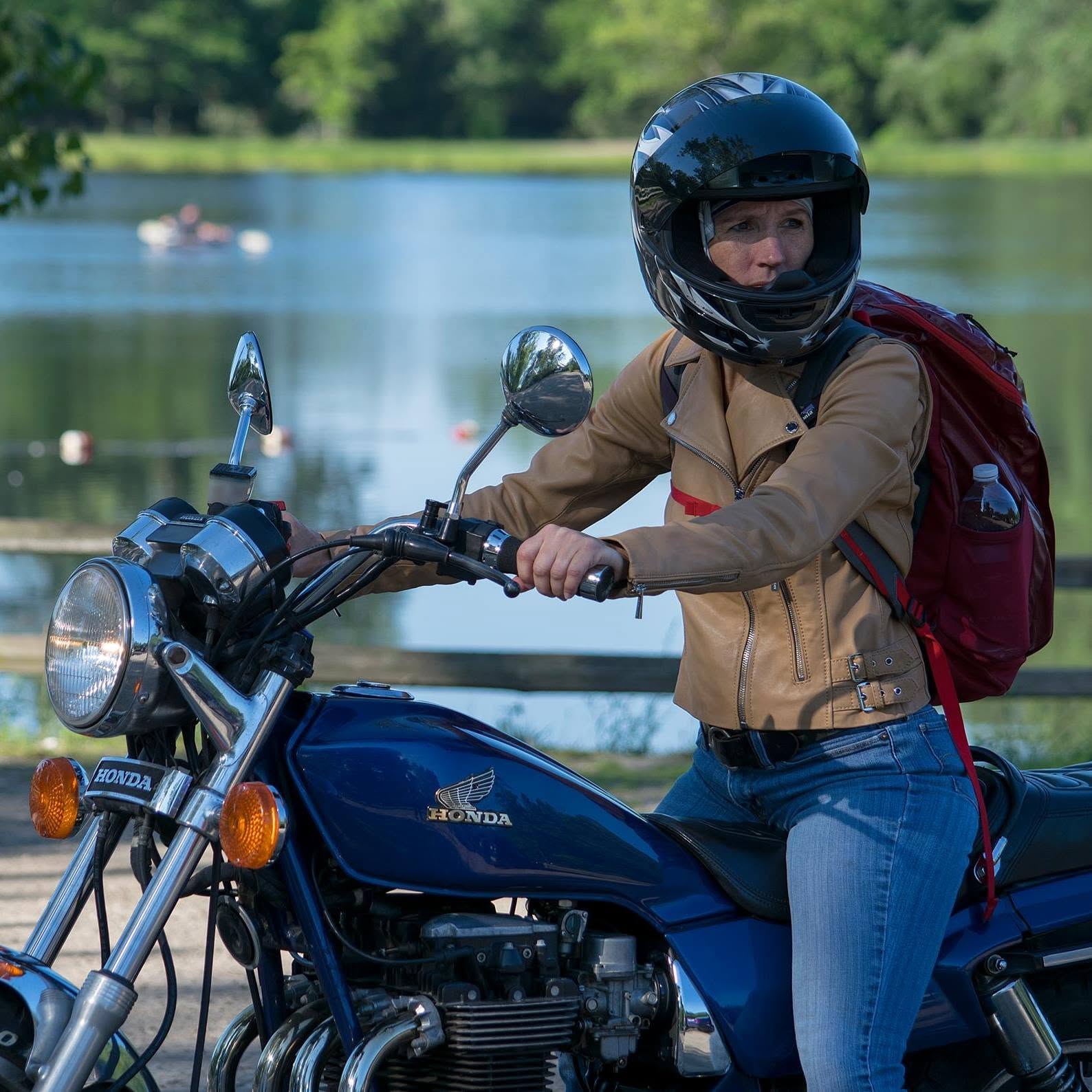 Person wearing a helmet and backpack sitting on a blue motorcycle near a lake with trees in the background.