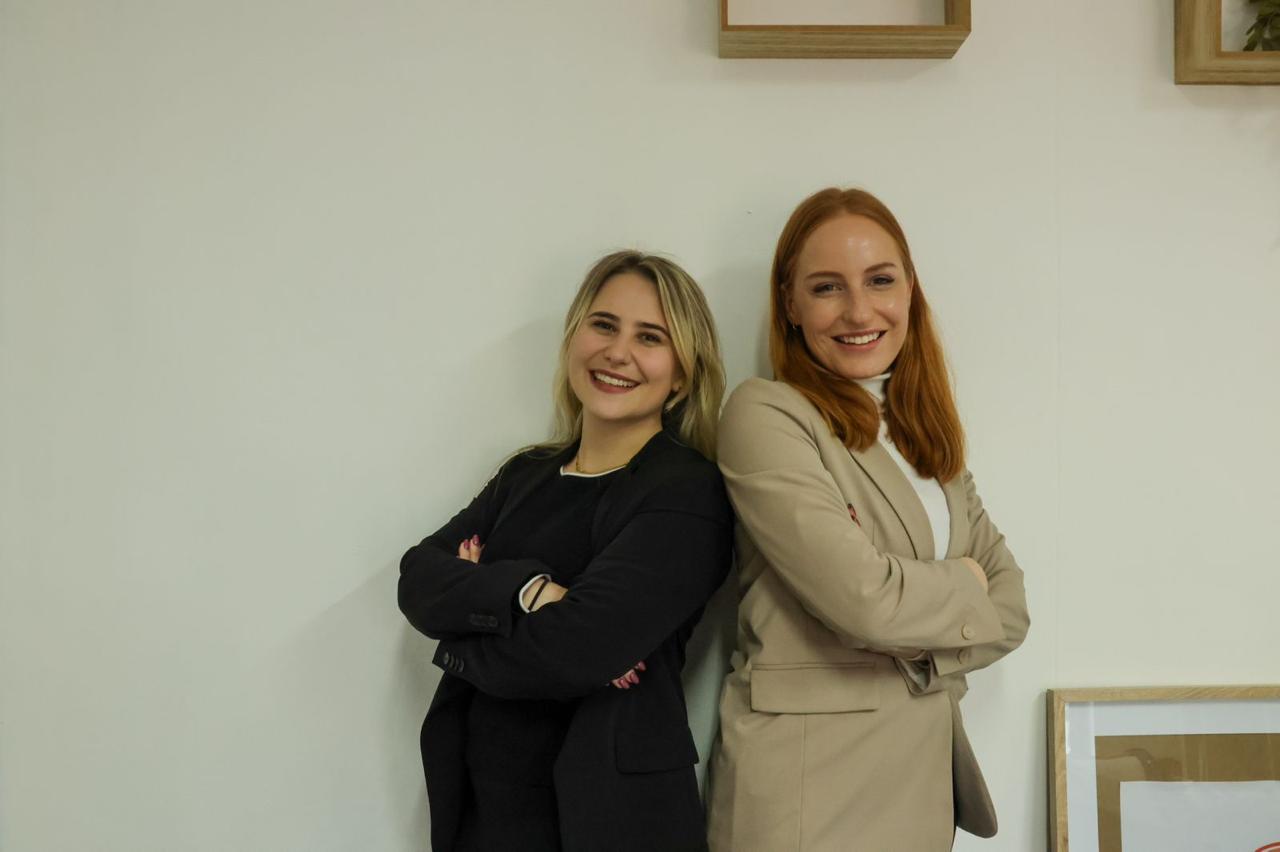 Two women standing side by side with arms crossed, smiling, against a plain wall with framed pictures.