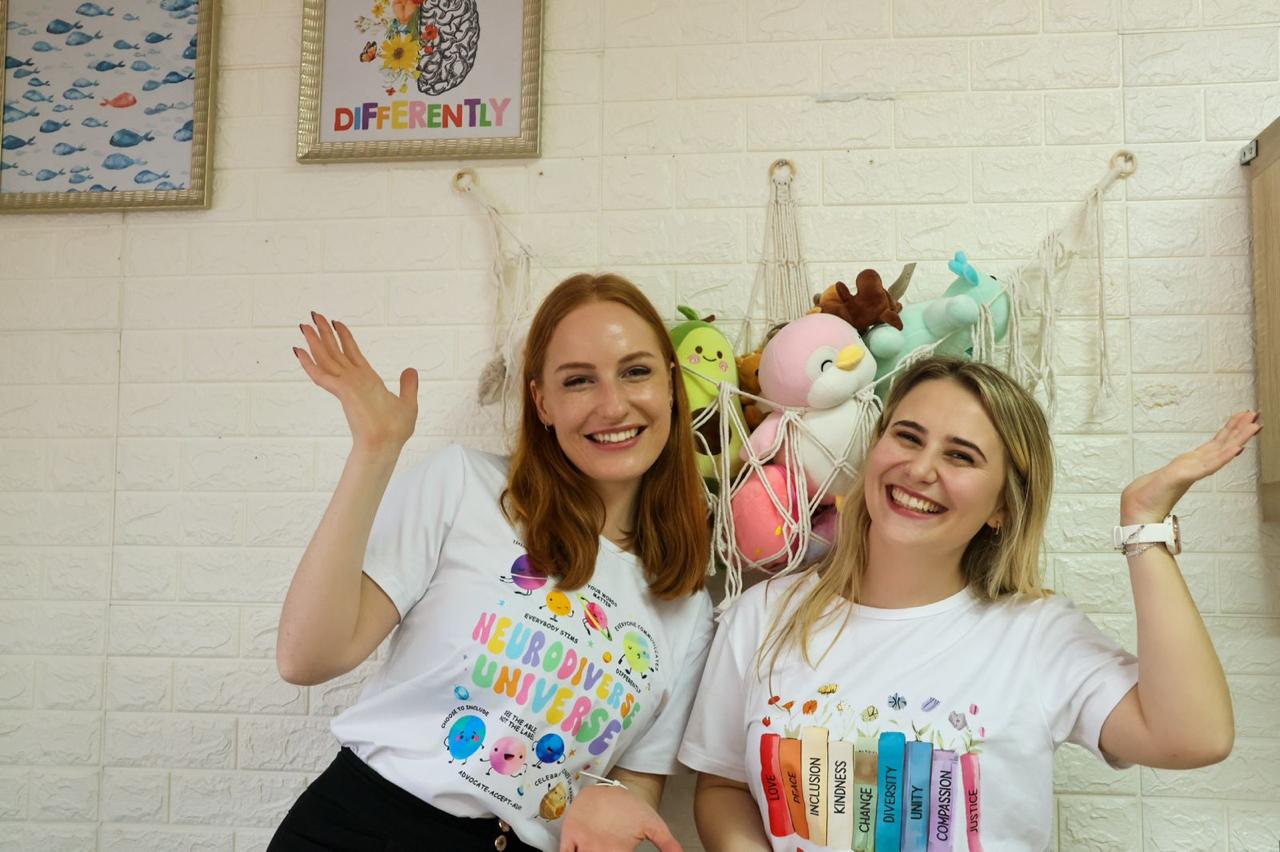 Two smiling women in white T-shirts with colorful designs, standing in front of a wall with plush toys and artwork.