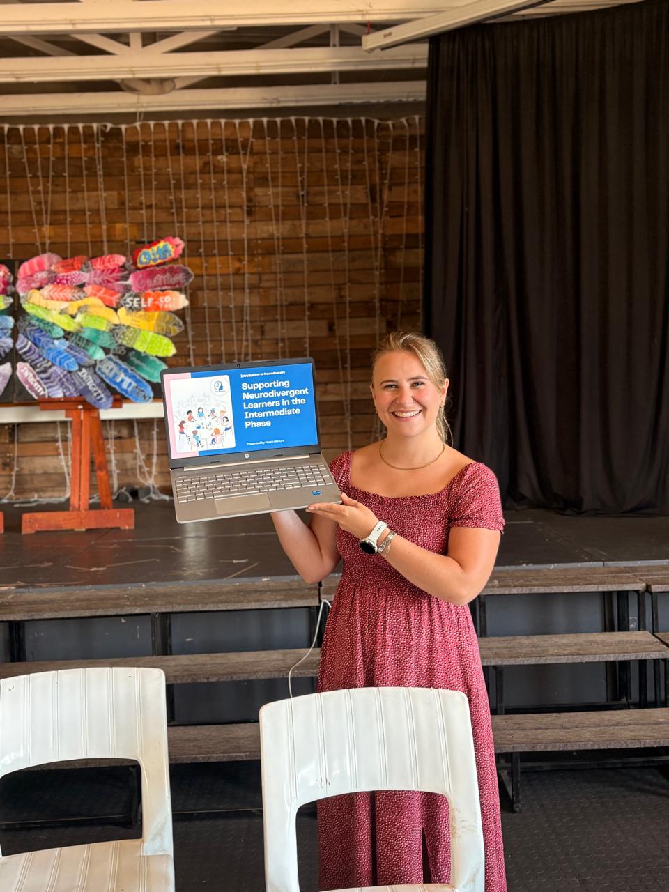 Smiling woman in a red dress holding a laptop displaying a presentation on stage with a colorful art piece in the background.