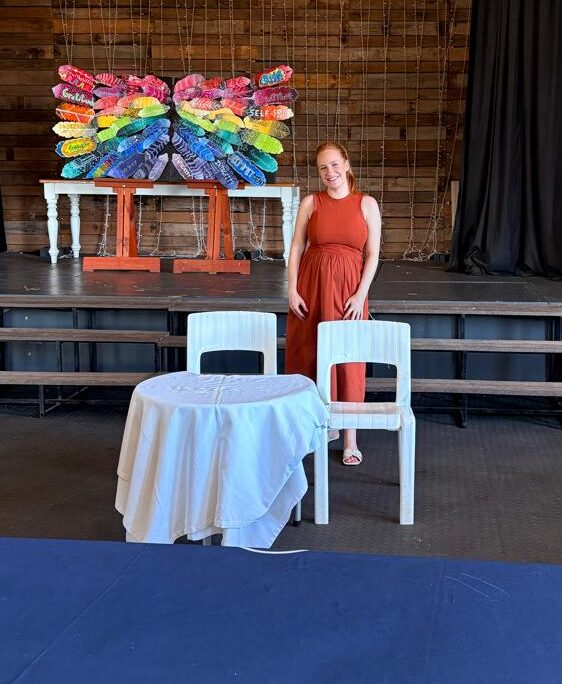 Woman in red dress standing behind a table with two chairs, colorful shoe display on stage behind her.