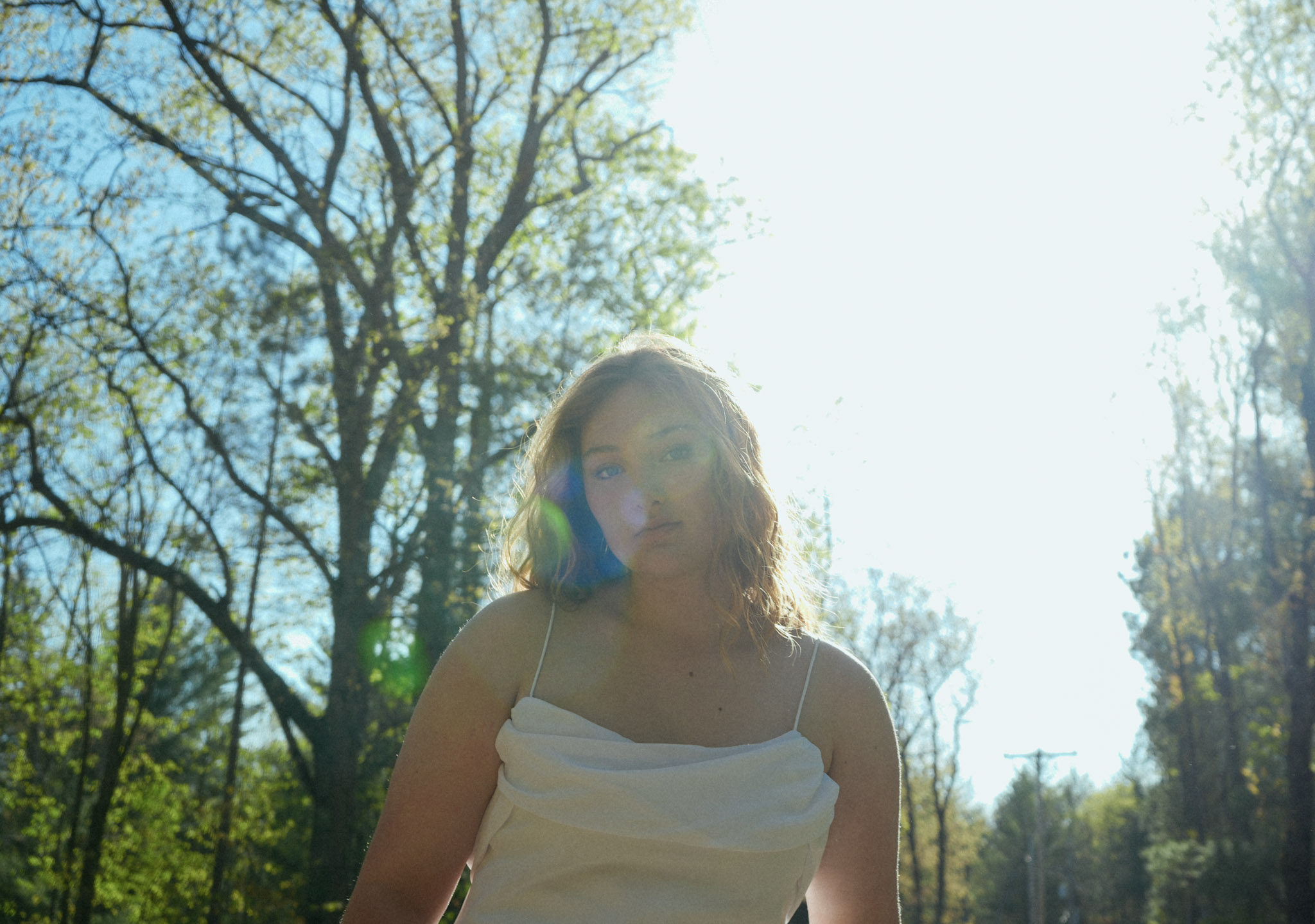 Young woman with wavy hair wearing a white top outdoors with trees and bright sunlight behind her.