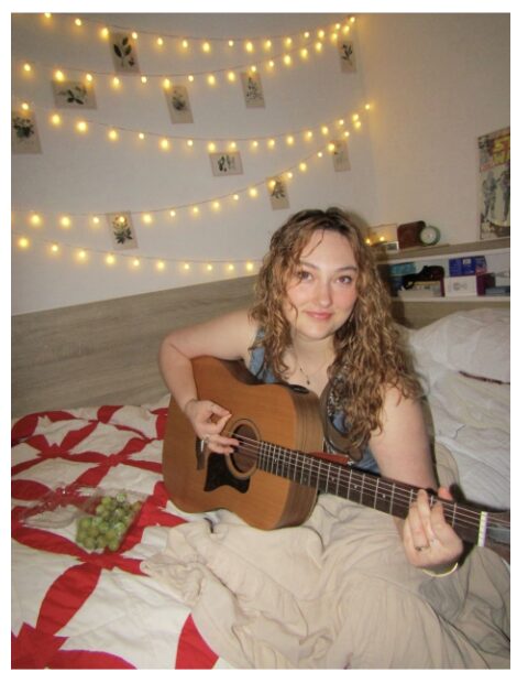 Young woman with curly hair sitting on bed, playing guitar, decorated with string lights and photos on wall.
