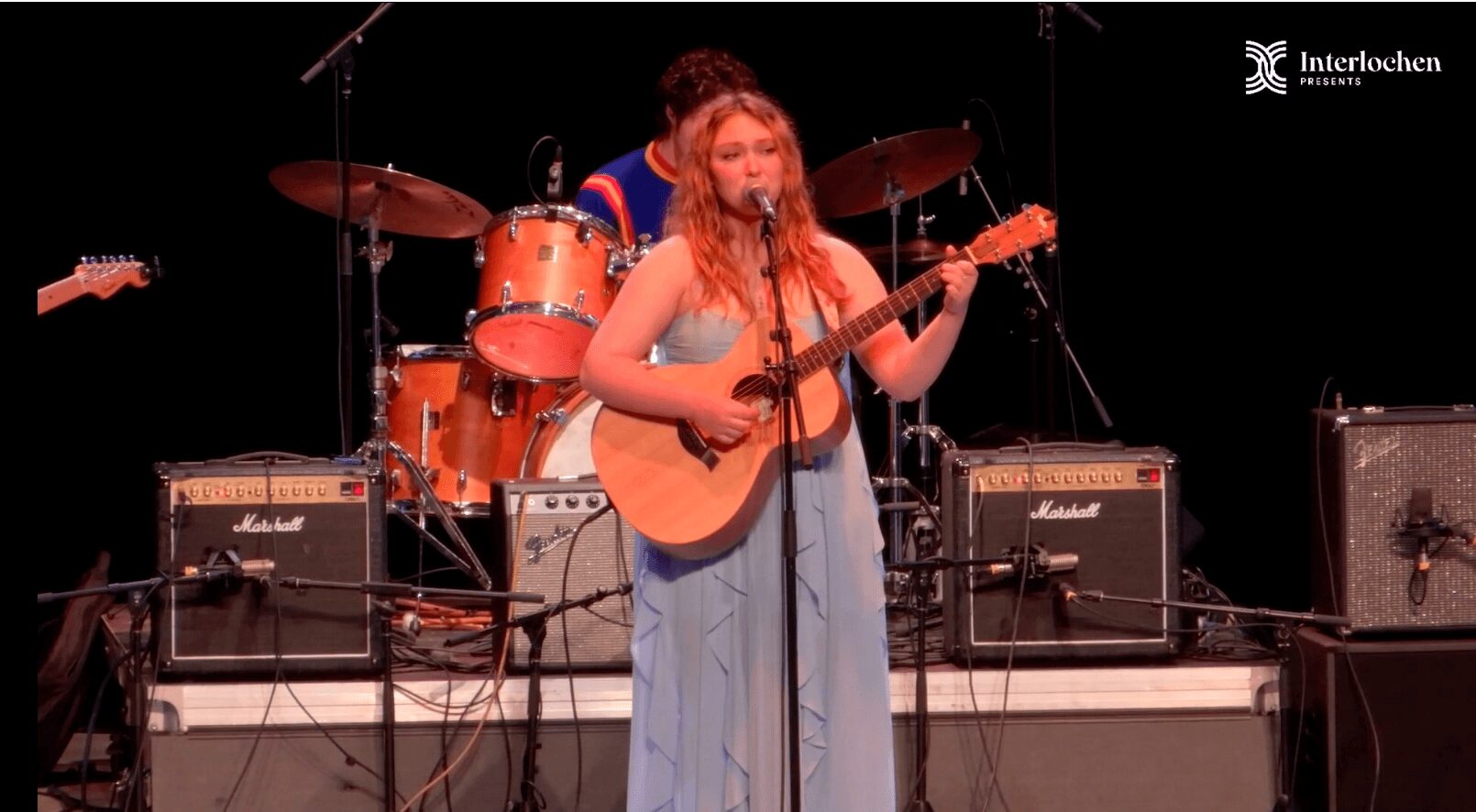 Woman playing guitar on stage with drum set and amplifiers behind her, performing in a concert setting.