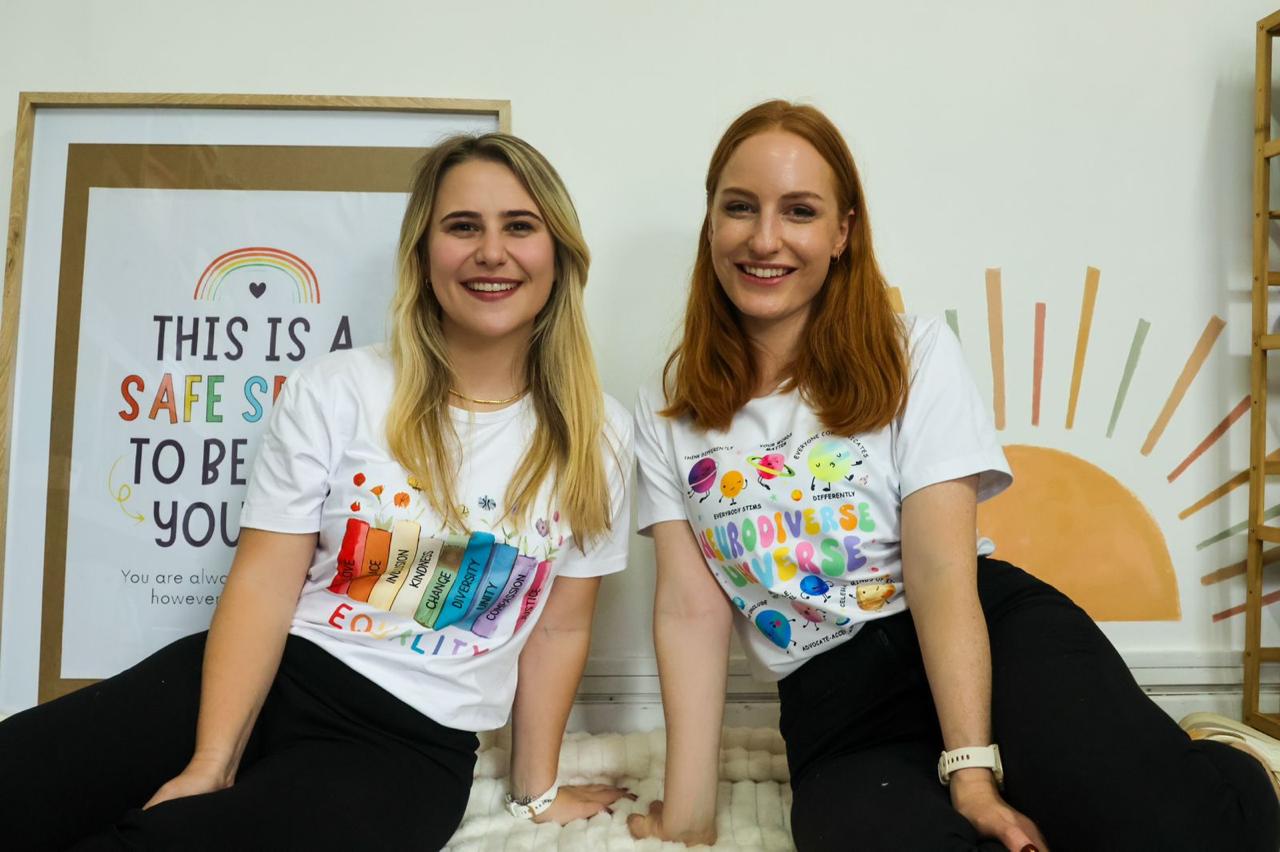 Two women sitting on the floor, smiling, wearing white T-shirts with colorful designs, in a bright room with a sun illustration and a sign. Word count: 20