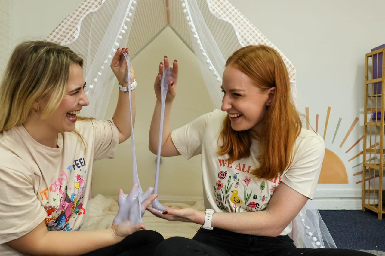 Two women smiling and playing with slime indoors, one holding slime and the other touching it, in a bright room.