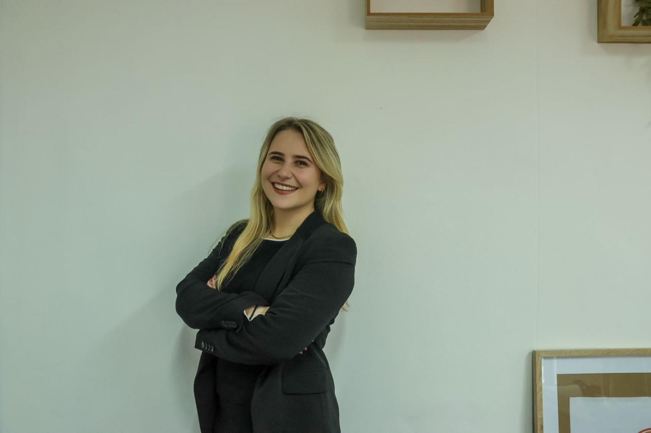 Young woman with long blonde hair smiling, standing with arms crossed against a plain wall.